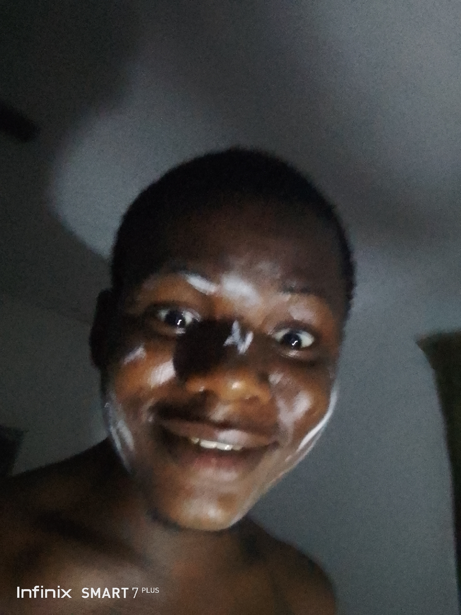 A person with dark skin is looking at the camera, with white cream on their face. The person is smiling, and their teeth are visible. The background is blurry and dark, but it appears to be indoors. The text "Infinix SMART 7 PLUS" is in the bottom left corner of the image.