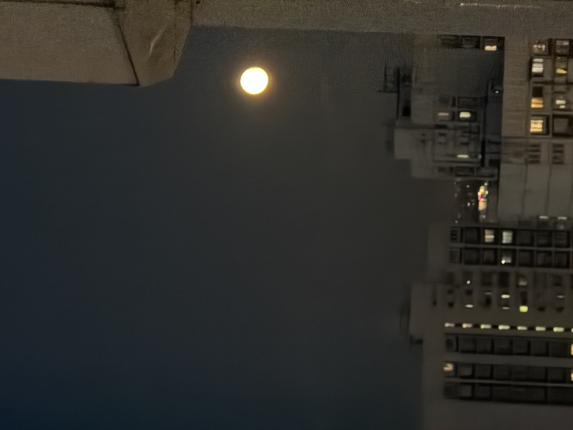 The image is a nighttime photograph taken from a high vantage point, likely a balcony or window, overlooking a city skyline. The sky is a dark, deep blue, with a bright, full moon positioned in the upper right quadrant of the image. The moon is a luminous yellow-white circle, emitting a soft glow. In the middle ground, the silhouettes of several multi-story apartment buildings are visible, with lights from many windows illuminating the facades. The buildings appear to be of uniform design, suggesting a residential area. The foreground on the right side of the image shows a textured, light-colored surface, possibly the exterior wall of the building from which the photo was taken, with vertical lines indicating construction joints or architectural features. The overall impression is a quiet urban night scene, with the moon dominating the sky above the illuminated buildings. The location is identified as Khimki, Russia.