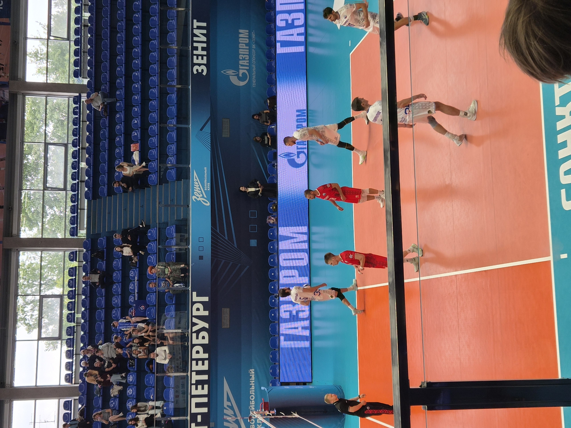 The image depicts a volleyball match taking place in an indoor arena. The court is bright orange with white lines marking the boundaries. Spectators are seated in blue tiered seating arranged around the court. The background features large windows overlooking trees and a bright sky. Sponsorship logos, including "Zenit" and "Gazprom," are visible on banners and the stadium walls. Players in red and white uniforms are on the court, some in action and others observing. The overall scene is indicative of a professional sporting event.