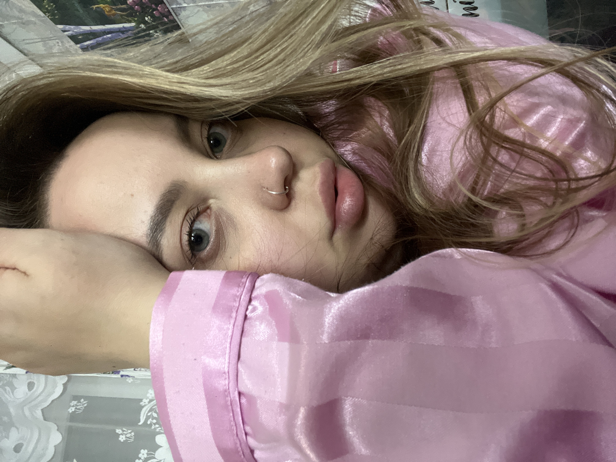 A blonde woman with a nose ring is lying down. She is wearing a pink satin pajama top. She is resting her head on her hand. The background includes a blurry image of what appears to be a landscape photograph and a white floral patterned surface. Location information is not discernible from the image.