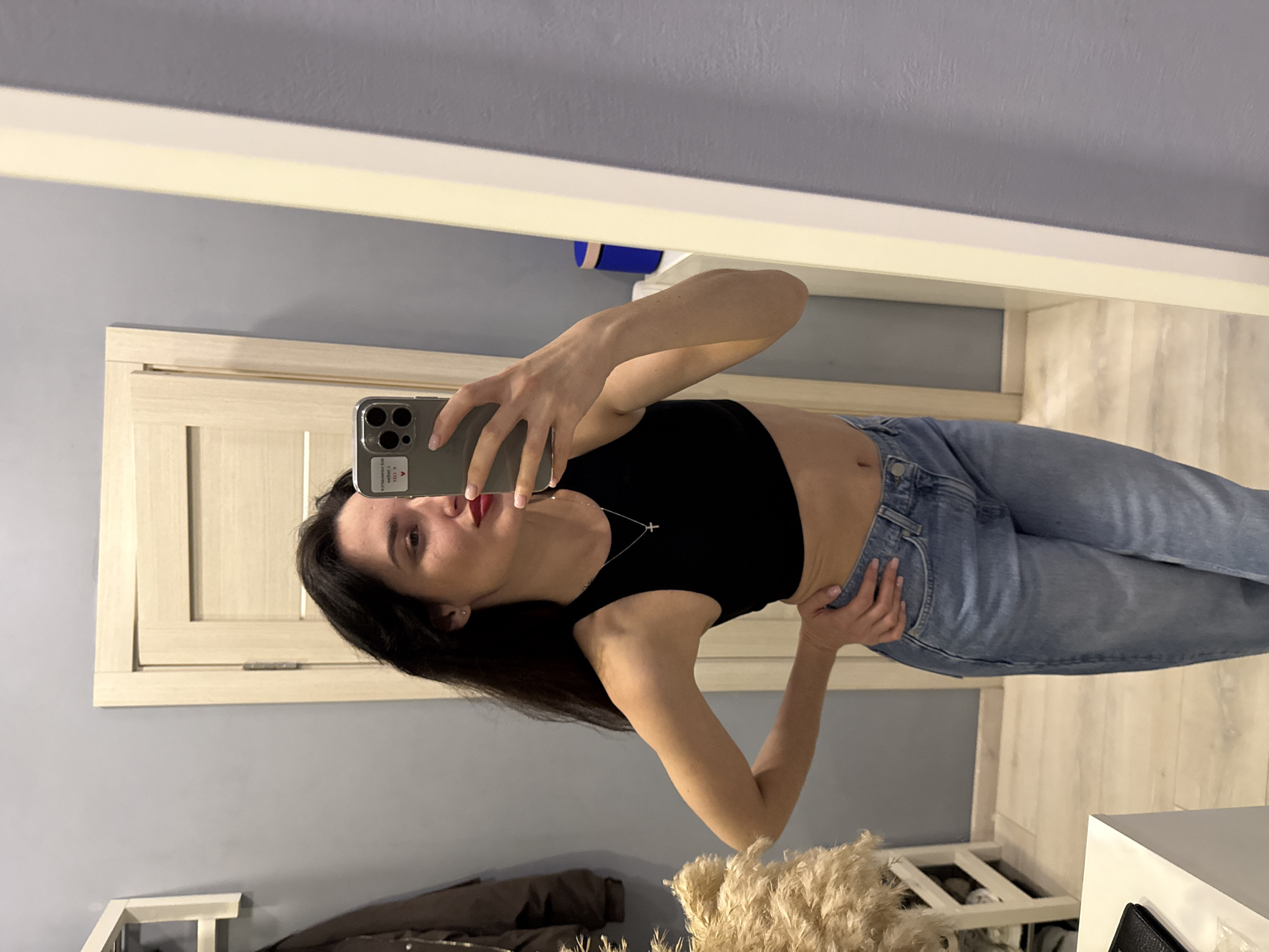 A woman is taking a mirror selfie indoors. She is wearing a black crop top and blue jeans. She is holding a phone with a sticker on it in her left hand. Her right hand rests on her hip. The background includes a mirror frame, a door, and a wall. The location is Unknown, Unknown.