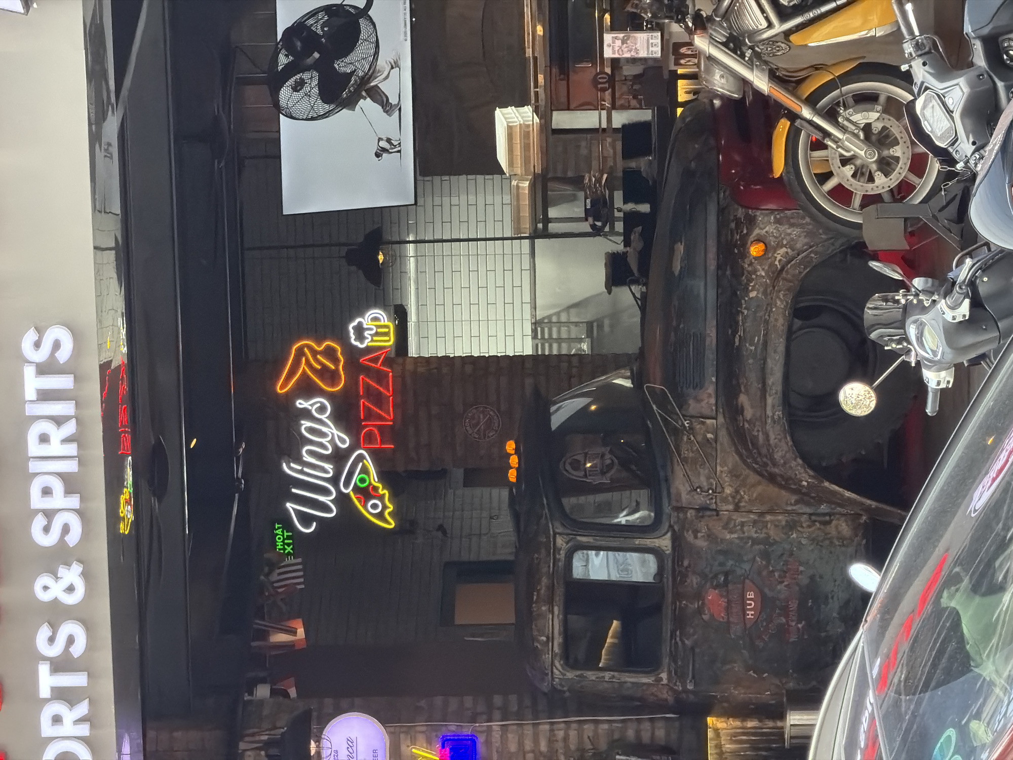 The image captures a vibrant, neon-lit street scene at what appears to be a bar or restaurant, likely in the evening or at night. The composition is dominated by various illuminated signs and objects.
In the center, a prominently displayed, rustic, dark brown truck cab, possibly an antique or repurposed vehicle, serves as a decorative element. The truck's surface shows significant wear and rust, contributing to an industrial or vintage aesthetic. On its side, faded red text reads "HUB."
Above and to the left of the truck, a bright orange and red neon sign spells out "Wings PIZZA," accompanied by an icon of a beer mug and a slice of pizza. Below this, smaller white text reads "THOÁT EXIT," indicating an exit and providing a strong clue to the Vietnamese location. Further to the left, running vertically along a building facade, another large white neon sign declares "SPORTS & SPIRITS."
Various motorcycles are visible in the upper right portion of the image, with one featuring a bright yellow fender and chrome details, parked or displayed alongside the truck. Other darker motorcycles are also partially seen, possibly reflected or parked nearby.
In the background, a white tiled wall is visible, suggesting interior or courtyard architecture. An industrial-style black fan hangs from the ceiling in the upper left, above a white poster or banner featuring a person. The overall impression is that of a lively establishment, combining food, drinks, and sports, with a distinct, perhaps retro or themed, decor.