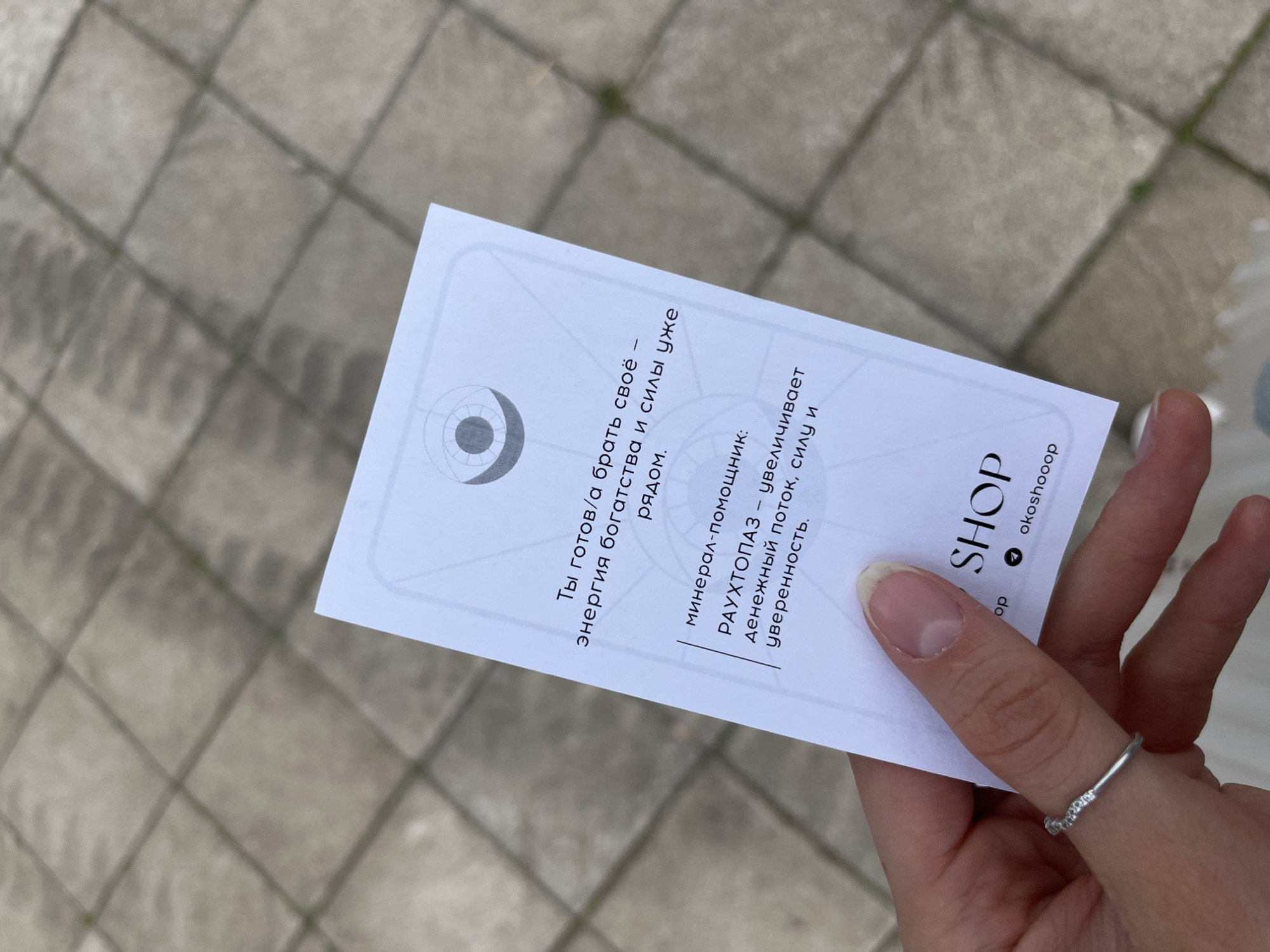 A hand holds a white card with text in Russian. The card has a symbol at the top and lists "РАУХТОПАЗ" (Rauchtopaz) as a mineral-helper that increases money flow, strength, and confidence. The card has the shop name "okoshooop". The background is a gray, tiled outdoor surface, possibly a sidewalk.