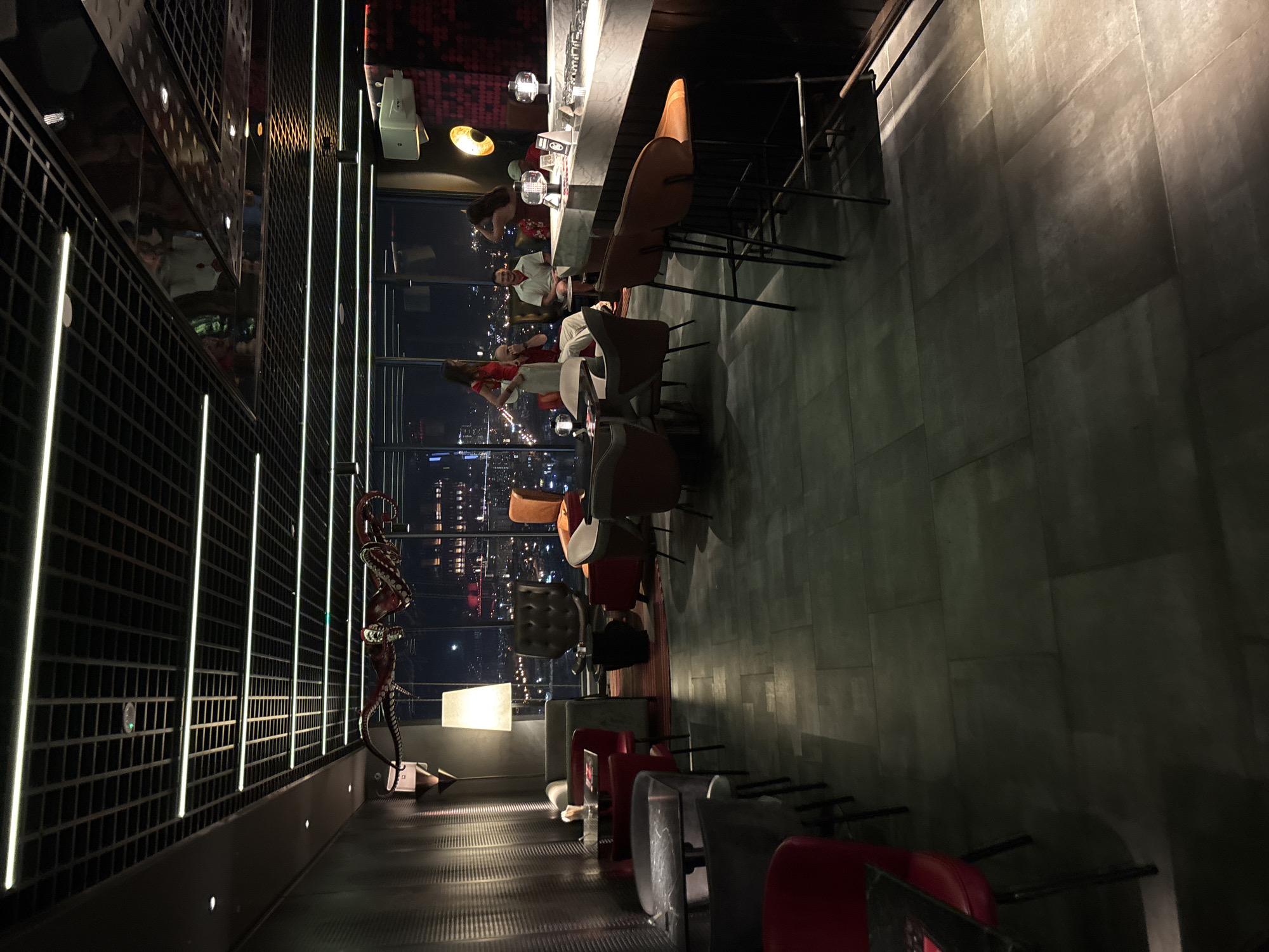 This media file depicts the interior of a modern, upscale bar or restaurant located in Đà Nẵng, Vietnam, viewed at night. The scene is characterized by its stylish design, featuring a long, polished bar counter with several stools, and a row of chairs and tables adjacent to large windows offering a nighttime city view.
The primary subjects are the people present and the bar's furnishings. Several individuals are visible, some seated at the bar and others standing or walking, engaged in conversations or enjoying the ambiance. The bar area is well-lit, with modern pendant lights and a bright counter surface. The chairs and tables are varied in design, contributing to a contemporary aesthetic. Notably, a decorative octopus sculpture is positioned along the wall, adding a unique artistic element. The background through the windows reveals the illuminated cityscape of Đà Nẵng at night, with numerous buildings and lights creating a vibrant urban panorama.
The time of day is clearly night, suggested by the dark sky and the illuminated city lights. The lighting within the bar is atmospheric, with accent lights along a patterned wall and soft illumination on the floor. The overall mood appears relaxed and social, consistent with a nighttime establishment.
The location is identified as Đà Nẵng, Vietnam, which is implied by the cityscape visible through the windows. The architecture and interior design suggest a sophisticated venue.
There is no visible text in the image that is legible or relevant to identifying specific details about the establishment or any events.
