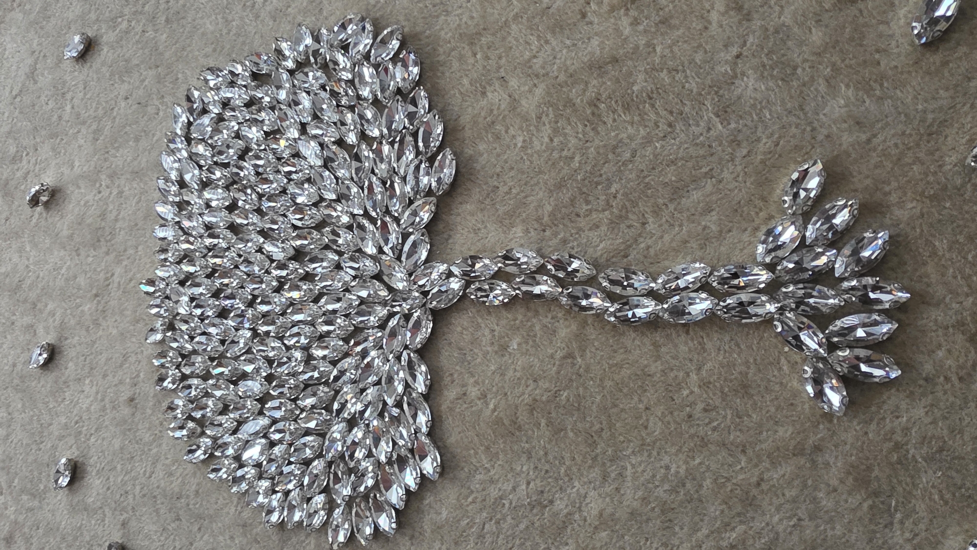 The image presents a close-up view of an ornate decorative pattern crafted from numerous clear, faceted, marquise-shaped rhinestones. These sparkling jewels are arranged on a soft, textured fabric surface, which appears to be a light brown or beige color, possibly felt or velvet.
The main part of the design forms a dense, fan-like or semi-circular cluster of rhinestones positioned on the left side of the frame. From this concentrated mass, a branching, vine-like element extends horizontally towards the right. This "stem" is composed of several segments, each made of pairs of rhinestones linked end-to-end. Further to the right, the stem bifurcates into smaller clusters of rhinestones, resembling stylized leaves or blossoms. A few individual, scattered rhinestones are also visible on the fabric, mainly to the left of the main pattern.
No activity or event is taking place; the image is a static shot of this intricate embellishment. The lighting suggests an indoor setting or diffused natural light, emphasizing the reflective qualities of the rhinestones. There are no visual cues within the image to indicate a specific time of day, weather conditions, or any particular emotions. Furthermore, no text is visible. While the location context of Blagoevgrad, Bulgaria, was provided, the image itself does not contain any geographical indicators.