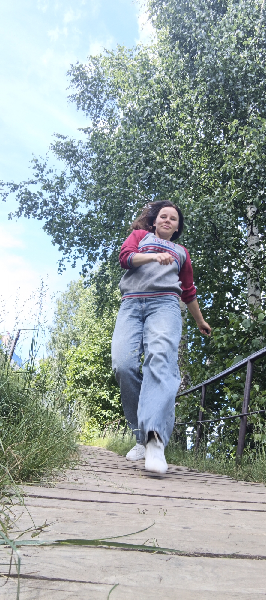The image depicts a young woman with dark hair, wearing a grey and red striped sweatshirt and loose-fitting blue jeans, as she walks down a wooden boardwalk. She is captured mid-stride, with her right leg slightly forward and her left arm extended to the side. The boardwalk, made of weathered wooden planks, is bordered by lush green grass and foliage. To the right of the boardwalk, a dark metal railing runs parallel to the path. Behind the woman and the boardwalk, a dense canopy of green trees, including what appears to be a birch tree with white bark, dominates the background, set against a bright blue sky with scattered white clouds. The scene appears to be outdoors in a natural setting, possibly a park or recreational area. While the specific location within Chaikovsky, Russia is not explicitly identifiable from the image, the overall environment suggests a locale with ample green spaces and wooden pathways common in such areas.