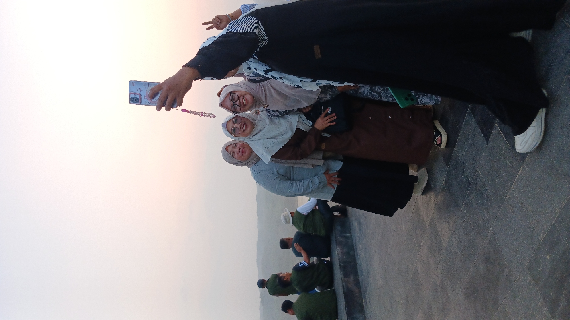 A group of four women wearing hijabs are taking a selfie outdoors. The woman on the far right is holding a phone and a string of beads is visible. The woman next to her is wearing glasses and a light-colored hijab and brown abaya. The third woman from the right is wearing a light blue hijab and a long black skirt. The woman on the far left is wearing a grey hijab and a blue shirt. In the background, several men are sitting on a ledge, facing away from the camera. The setting appears to be a scenic overlook, with rolling hills and a body of water in the distance. The sky is a pale orange, suggesting either sunrise or sunset. The ground is paved with dark grey tiles. The location is identified as Tanjungsari, Indonesia.