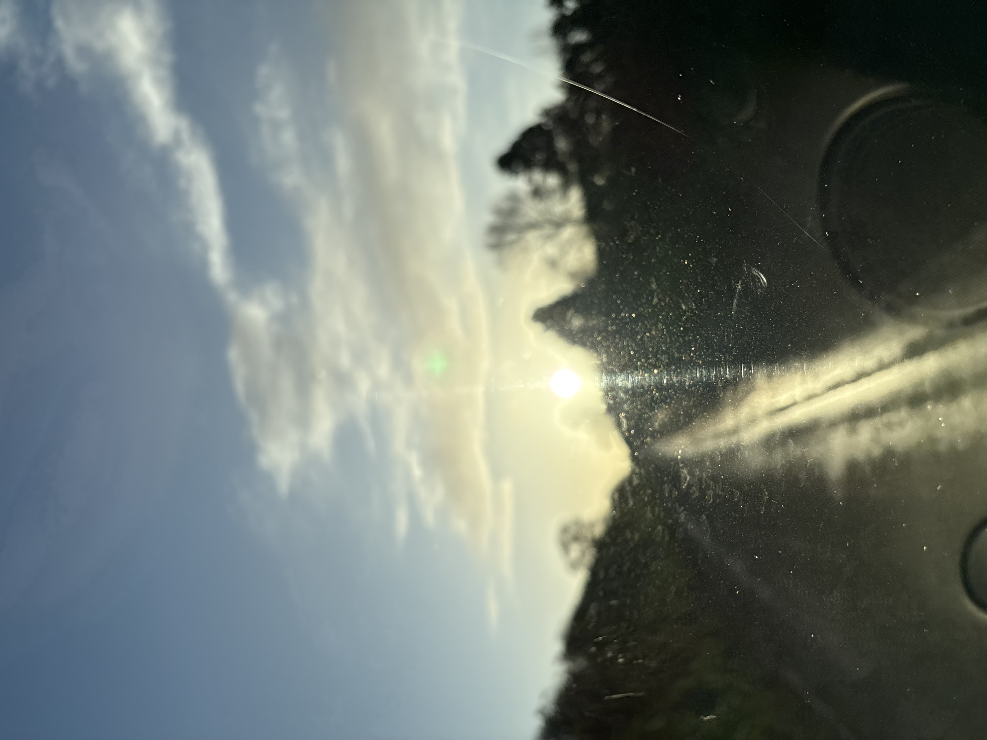 The image captures an exterior scene from inside a vehicle, viewed through a dirty or wet windshield, located in Ruyton-XI-Towns, United Kingdom. A prominent, low-lying sun is positioned near the center of the horizon, obscured and causing significant glare and lens flare, including a distinct green artifact above and to its left. Numerous illuminated dust particles, streaks, and potential water droplets are visible on the windshield surface, particularly in the lower half. A diagonal scratch or streak traverses the lower right quadrant of the windshield. The sky above is partly cloudy, displaying shades of blue, white, and light grey, with clouds illuminated by the low sun. The landscape, consisting of silhouetted trees and hills, is heavily blurred and appears as dark masses against the horizon. A road surface, highly reflective due to the sun's angle, extends forward from the vehicle, exhibiting two distinct parallel streaks of reflected light. No individuals or specific actions are discernable.
