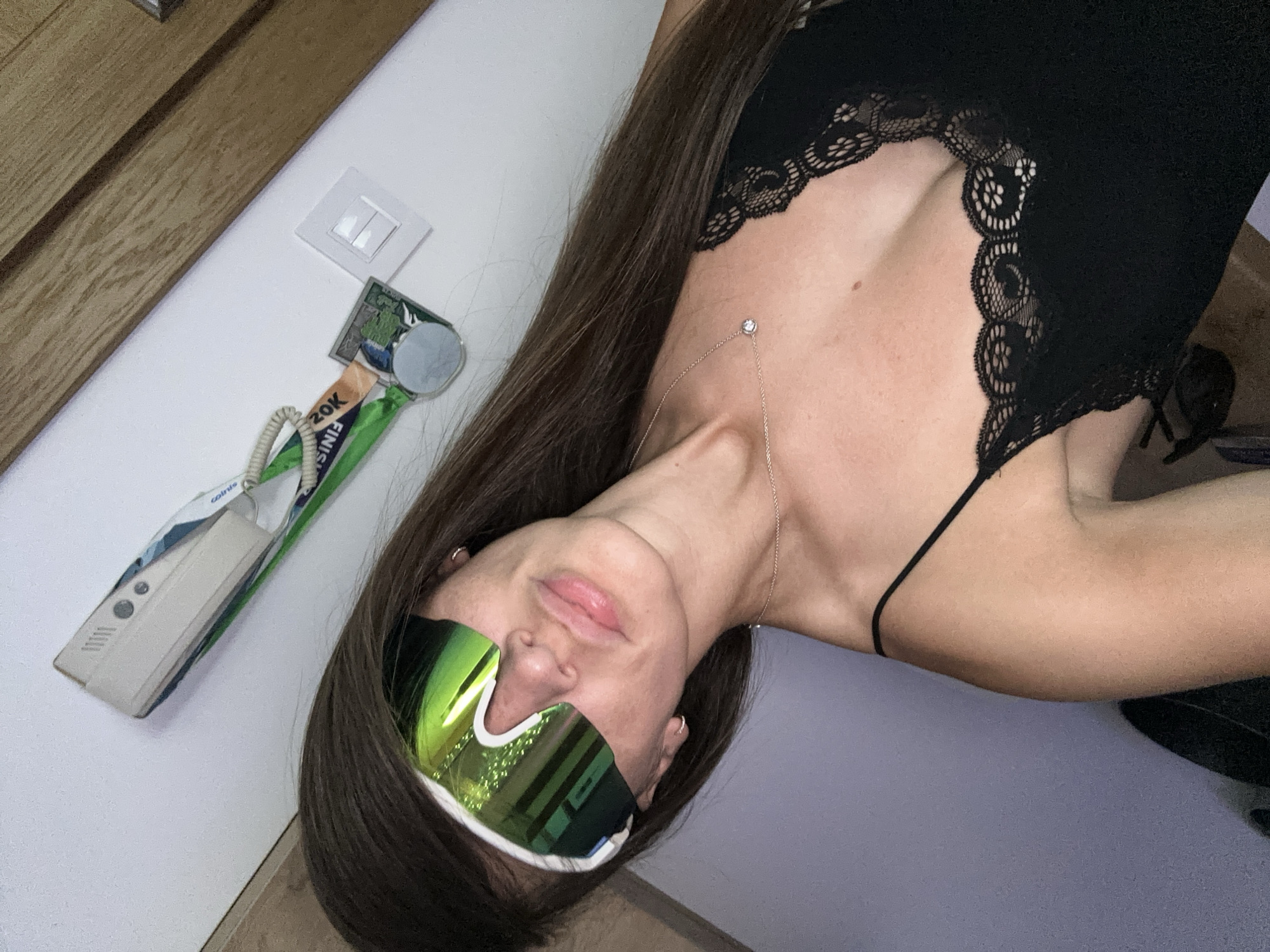 The media file shows a woman lying down, with her head tilted. She is wearing a black lacy camisole and reflective green goggles. A thin gold chain with a small pendant is visible around her neck. Her long, dark hair is spread out.
To her left, on a white surface, are several objects including a beige intercom device with a coiled cord, and what appear to be race medals or ribbons, one of which has text partially visible that looks like "20K FINIS". A light switch is also visible on the white wall. A wooden element, possibly a door or furniture, is to the left of the frame.
The scene appears to be indoors, with artificial lighting. The overall impression is casual and relaxed. The woman's pose and the presence of race medals suggest she might be resting after a sporting event. The image is oriented such that the woman's head is at the bottom of the frame. The background is a plain light purple wall. No specific time of day or weather conditions can be determined.