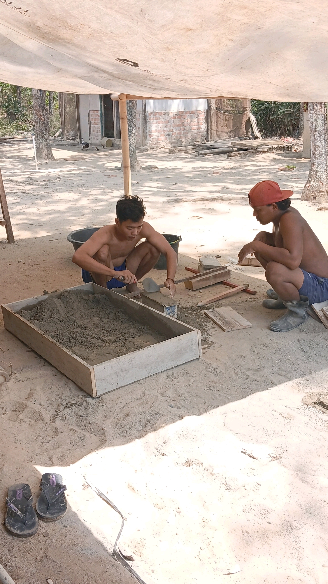 The image depicts two young men in Adi Luhur, Indonesia, engaged in what appears to be a construction-related activity under a makeshift canvas shelter. Both individuals are squatting on a sandy, dirt ground. The man on the left, wearing blue shorts, is focused on a wooden frame filled with a grey, granular material resembling cement or sand. He is using a small trowel to manipulate the material within the frame. A dark plastic tub is situated behind him.
The man on the right, wearing blue shorts and an orange baseball cap, is also squatting and observing the man on the left. He is wearing dark boots. Several wooden planks and blocks are scattered around them on the ground, along with some tools. In the background, there is a partially constructed brick building, some trees, and vegetation, indicating an outdoor setting in a semi-rural or developing area. A pair of dark flip-flops are visible in the lower left corner of the frame. The overall lighting suggests it is daytime, with strong shadows cast by the shelter and objects.
