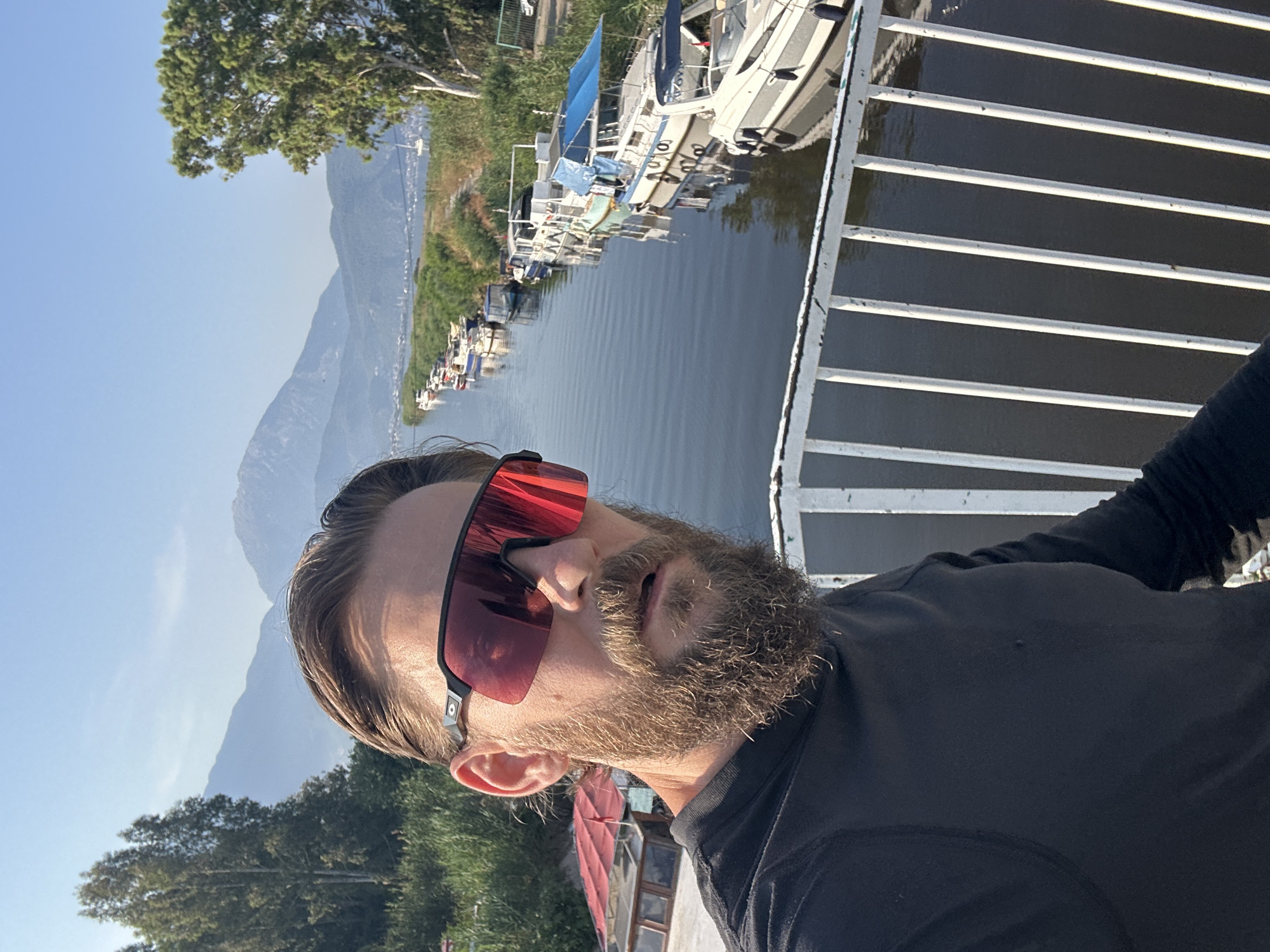 The image is a selfie taken by a man with a beard and sunglasses, standing on a white metal railing. He is wearing a black long-sleeved shirt. The background shows a canal with several boats docked along its banks. The canal is lined with green vegetation, and in the distance, there are mountains under a clear blue sky. The scene is likely located in Fethiye, Turkey, possibly in an area with canals and boat docks, such as the Fethiye marina or a nearby waterway. The sunlight suggests it is daytime.