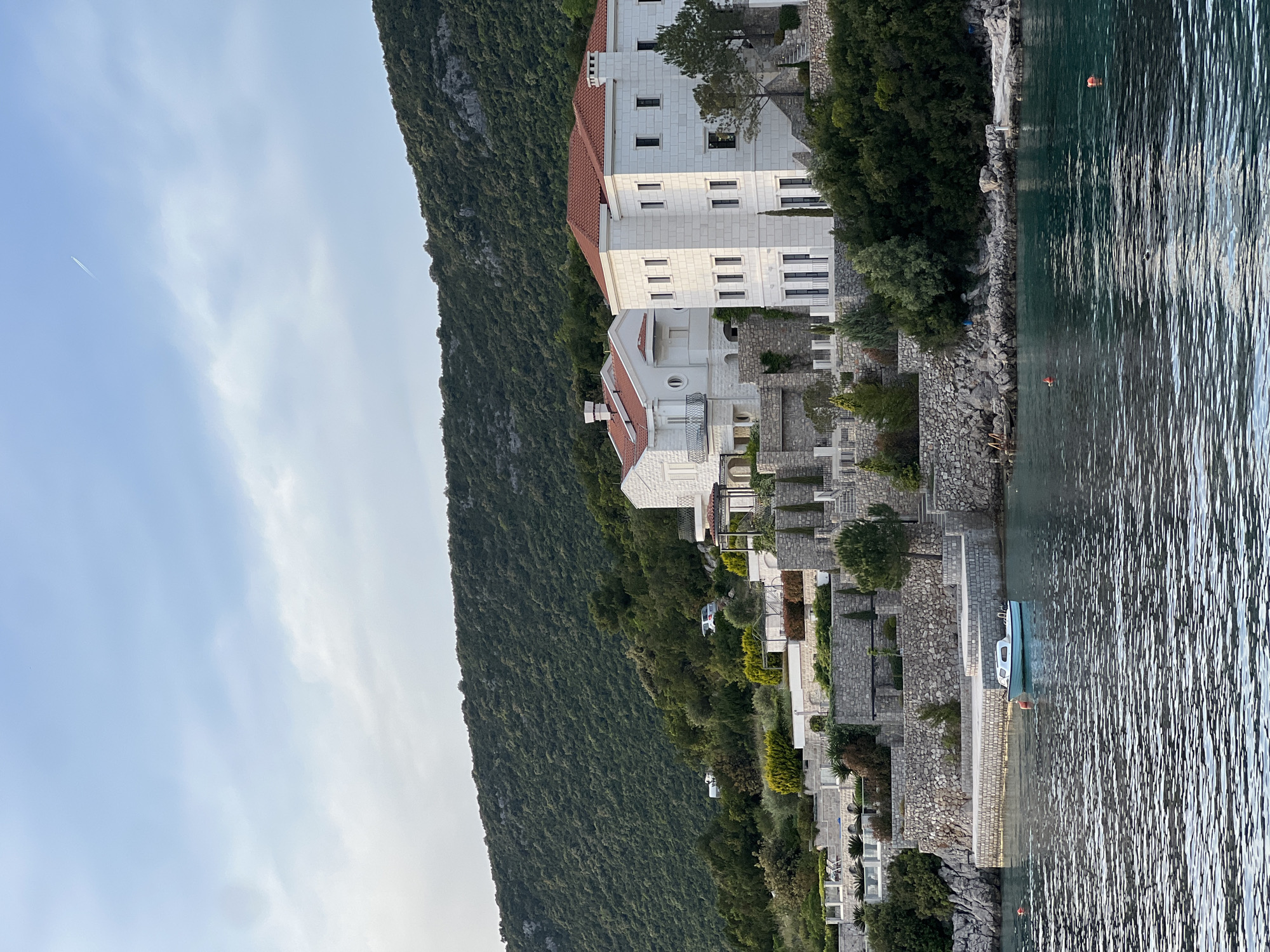 The image shows a view of the coast in Luštica, Montenegro. A mountain covered in green vegetation rises on the left. A multi-story white building with a red tile roof is built into the slope. Adjacent to the building are stone structures and vegetation leading down to the water's edge. A small boat is docked at a stone pier. The water is calm with visible ripples. The sky above the mountain is pale blue with light clouds. A contrail from an airplane is visible in the sky.