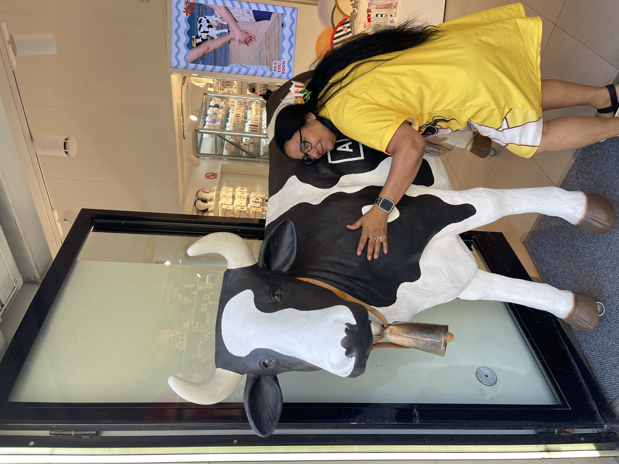 In Barcelona, Spain, a woman in a bright yellow dress is embracing a large, black and white cow statue outside a shop. The cow statue is detailed with horns, a bell around its neck, and realistic eyes. The woman has on glasses and her dark hair is long and flowing. The shop appears to be a retail establishment, with shelves of merchandise visible through a glass door. A poster featuring a cow and the text "SEA YOU SOON" is displayed to the right of the cow statue. The ground is covered with a dark gray mat.