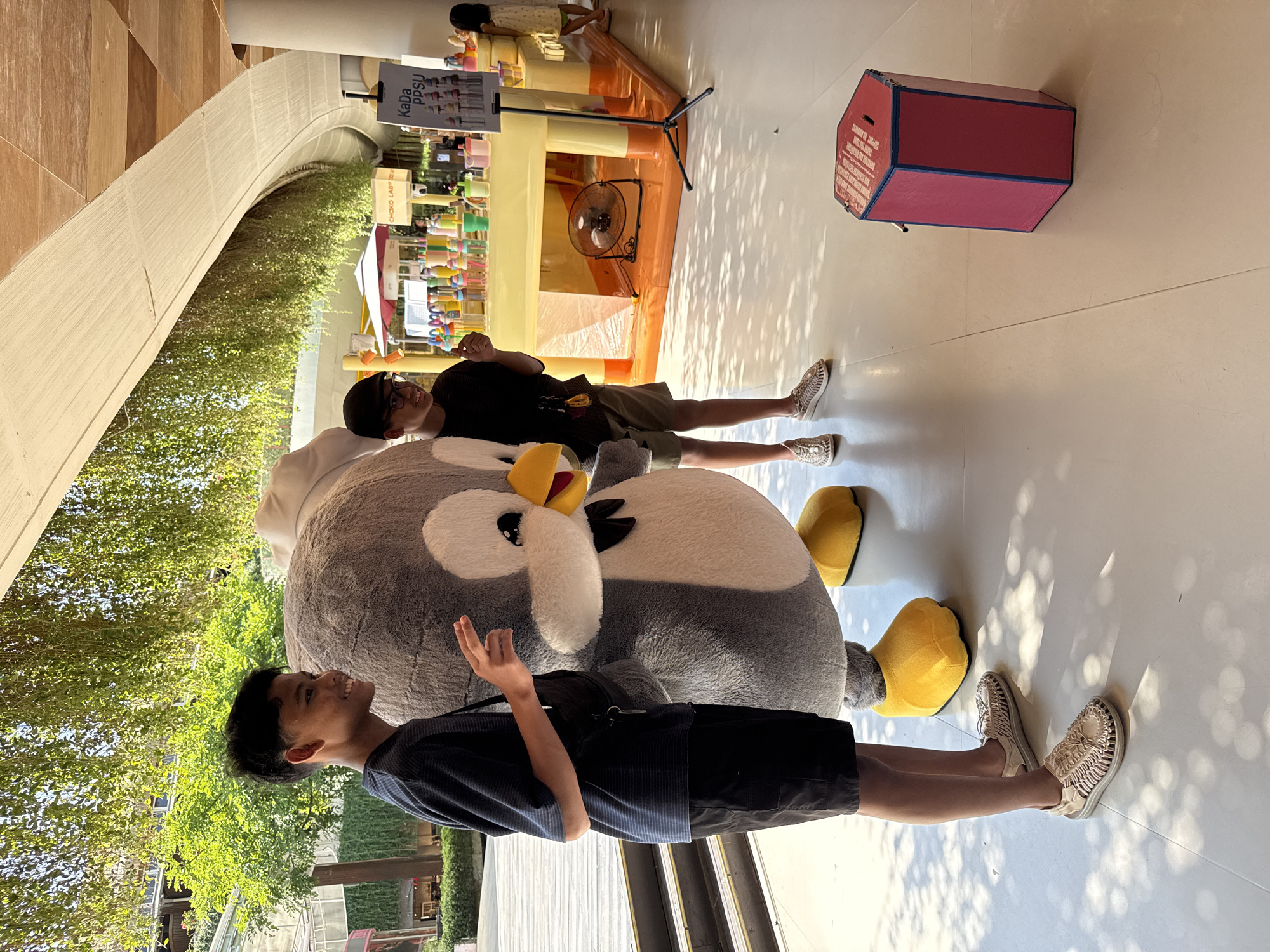 The image depicts two young males standing next to a large, plush penguin mascot. The male on the left wears a dark blue striped t-shirt and black shorts, with a small black bag slung across his chest. The male on the right wears a black t-shirt and olive green shorts, with glasses and a black cap. Both individuals are smiling and appear to be interacting with the mascot. The mascot is grey with a white chest and belly, a yellow beak, and black bow tie, and is wearing a white chef's hat.
Behind the individuals and mascot is a bright yellow structure with various colorful items displayed, resembling a food stall or play area. There are rows of colorful ice cream cones or similar items on display, along with a sign that reads "KaDa PPSU." A small fan is visible beneath the yellow structure.
The environment appears to be an indoor or covered outdoor space with a light-colored tiled floor and a wooden-paneled ceiling. Sunlight streams in, creating shadows on the floor. Green foliage hangs from above, suggesting an open or garden-like setting. In the foreground, to the right, is a pink hexagonal box with blue trim, possibly a donation or collection box. The scene suggests a family-friendly venue in Kuta, Indonesia, possibly a shopping mall or entertainment complex, given the architectural style and the presence of commercial activities.