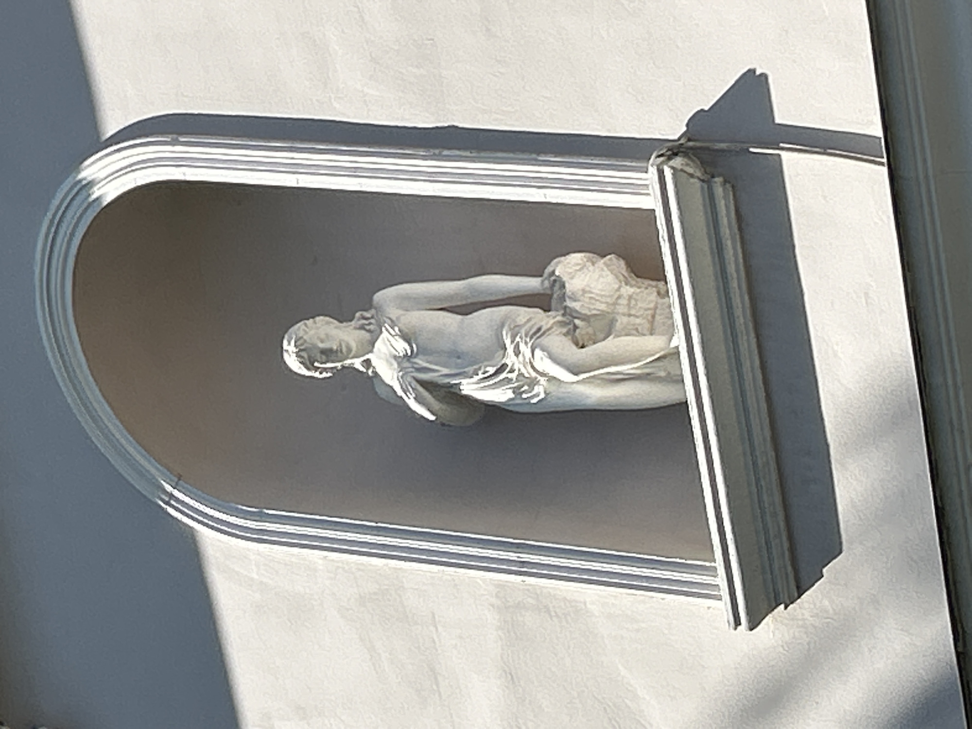 The image shows a white neoclassical statue of a woman in a niche on the facade of a building. The statue depicts a draped female figure with her left arm resting on a rock. The niche is arched and decorated with fluted moldings. The building's facade is painted a light color and is illuminated by sunlight, casting shadows. This architectural detail is located on the building at 7 Tverskaya Street in Moscow, Russia, which houses the Central Children's Store.