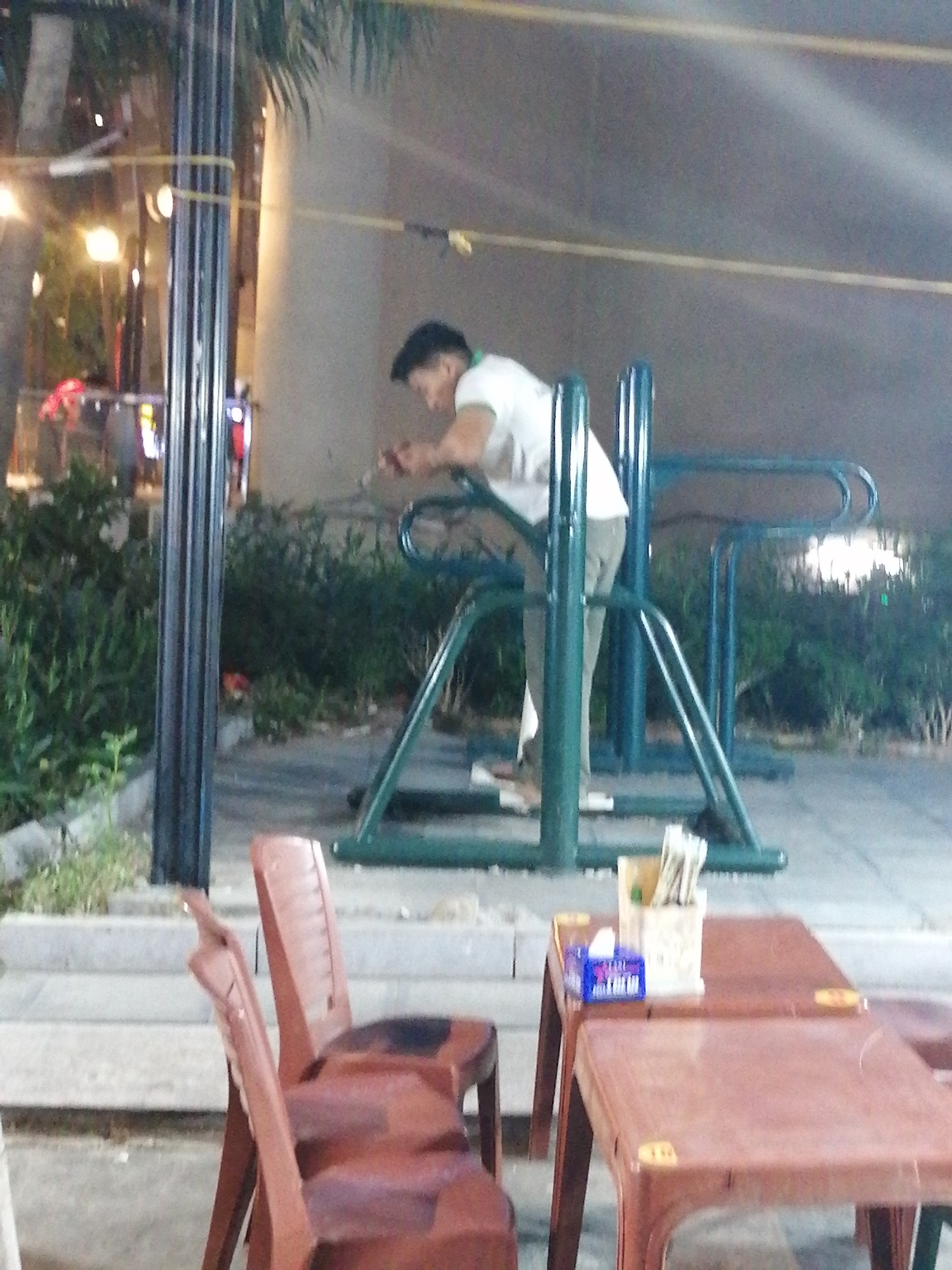 The image depicts an outdoor scene in Bai Chay Ward, Vietnam. A man in a light-colored t-shirt and pants stands near a green metal bike rack, seemingly looking at a device in his hands. In the foreground, there are two brown plastic chairs positioned in front of a brown plastic table. On the table, there is a tissue box and a container holding utensils. A black pole is situated to the left of the bike rack. Green bushes are visible behind the rack. In the background, a building and faint street lights are discernible.