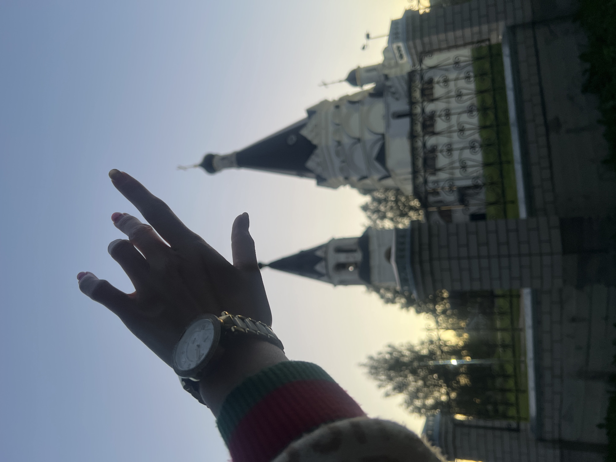 In this low-angle shot, a person's hand, adorned with a watch and painted nails, is raised towards a clear sky. The hand is silhouetted against the light, obscuring the person's face but revealing a portion of their arm clad in a sweater with red and green stripes. In the background, a white church with dark spires and intricate architectural details stands prominently. The church is partially obscured by trees and a decorative metal fence. The lighting suggests it is either early morning or late afternoon, with a soft, warm glow illuminating the scene. The overall mood is serene and contemplative, with the hand reaching towards the sky and the majestic church serving as a backdrop. The setting is likely Krasnoe-na-Volge, Russia, given the characteristic architecture of Russian Orthodox churches.