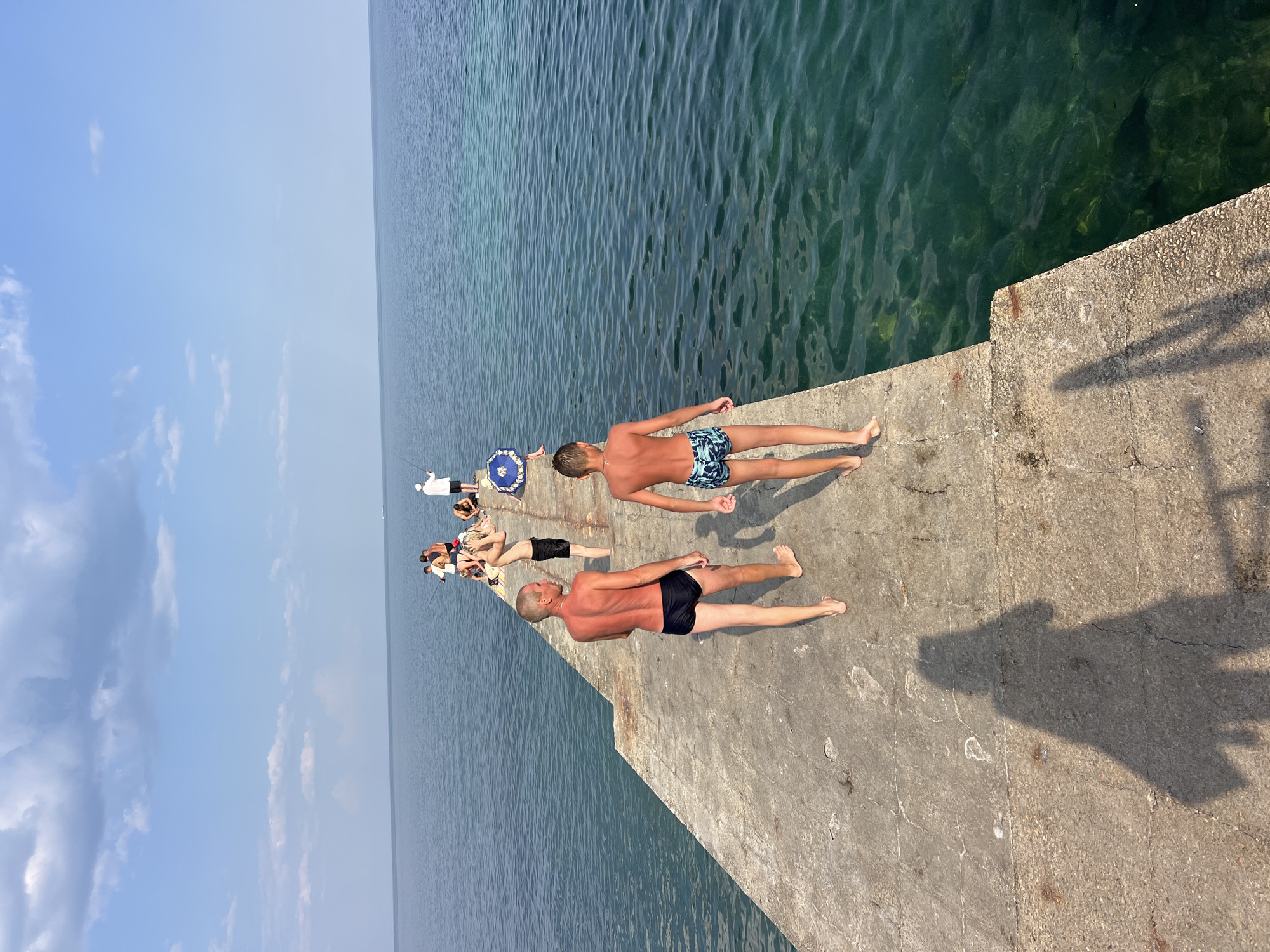 The image depicts a concrete pier extending into a clear, turquoise sea under a bright, cloudy sky. Several individuals are present on the pier. In the foreground, two males, an adult and a younger boy, are walking away from the camera, both wearing swim trunks. The adult has a tanned physique. Further down the pier, a group of people are scattered, some appearing to be fishing with rods, while others are seated or standing. A blue umbrella is visible among the group. The water is calm with visible ripples, and the sunlight casts shadows on the pier, including a prominent shadow of the photographer. The location is a coastal area, consistent with Odesa, Ukraine, known for its piers and seaside activities.