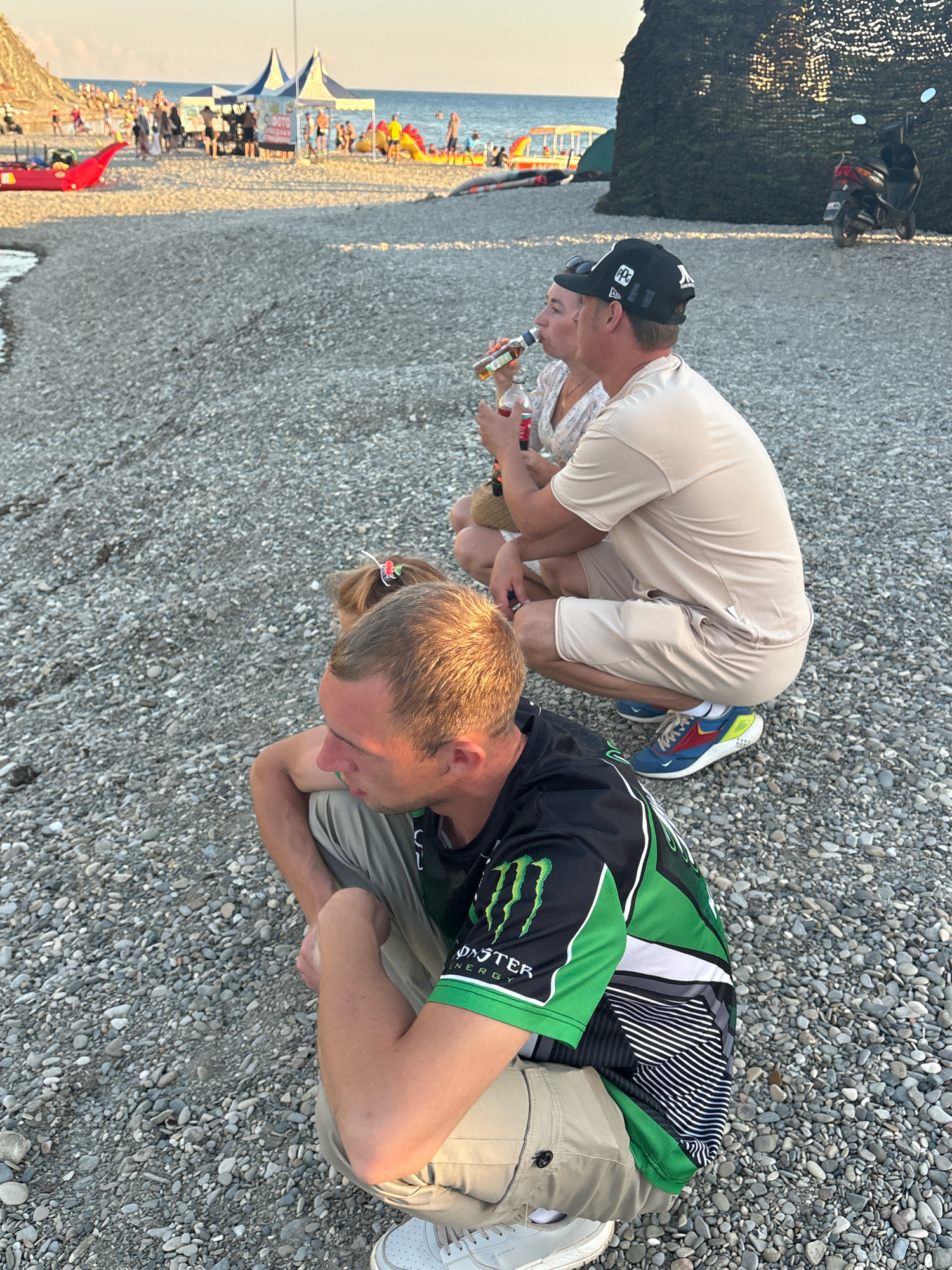 In Arkhipo-Osipovka, Russia, on a pebble beach, three individuals are crouched down. In the foreground, a man wearing a green and black Monster Energy shirt and tan shorts is facing to the right, with his left arm resting on his knee. Behind him and slightly to the left, a woman with blonde hair tied back with a red accessory is also crouched, facing left and drinking from a bottle. To her right, a man wearing a tan t-shirt and shorts, and a black baseball cap, is crouched and looking towards the left, also holding a bottle. The beach is covered in grey pebbles, and the sea is visible in the background, with several beach umbrellas, people, and inflatable attractions on the sand. A scooter is parked on the right side of the image, behind a camouflage-netted structure. The lighting suggests it is late afternoon or early evening.