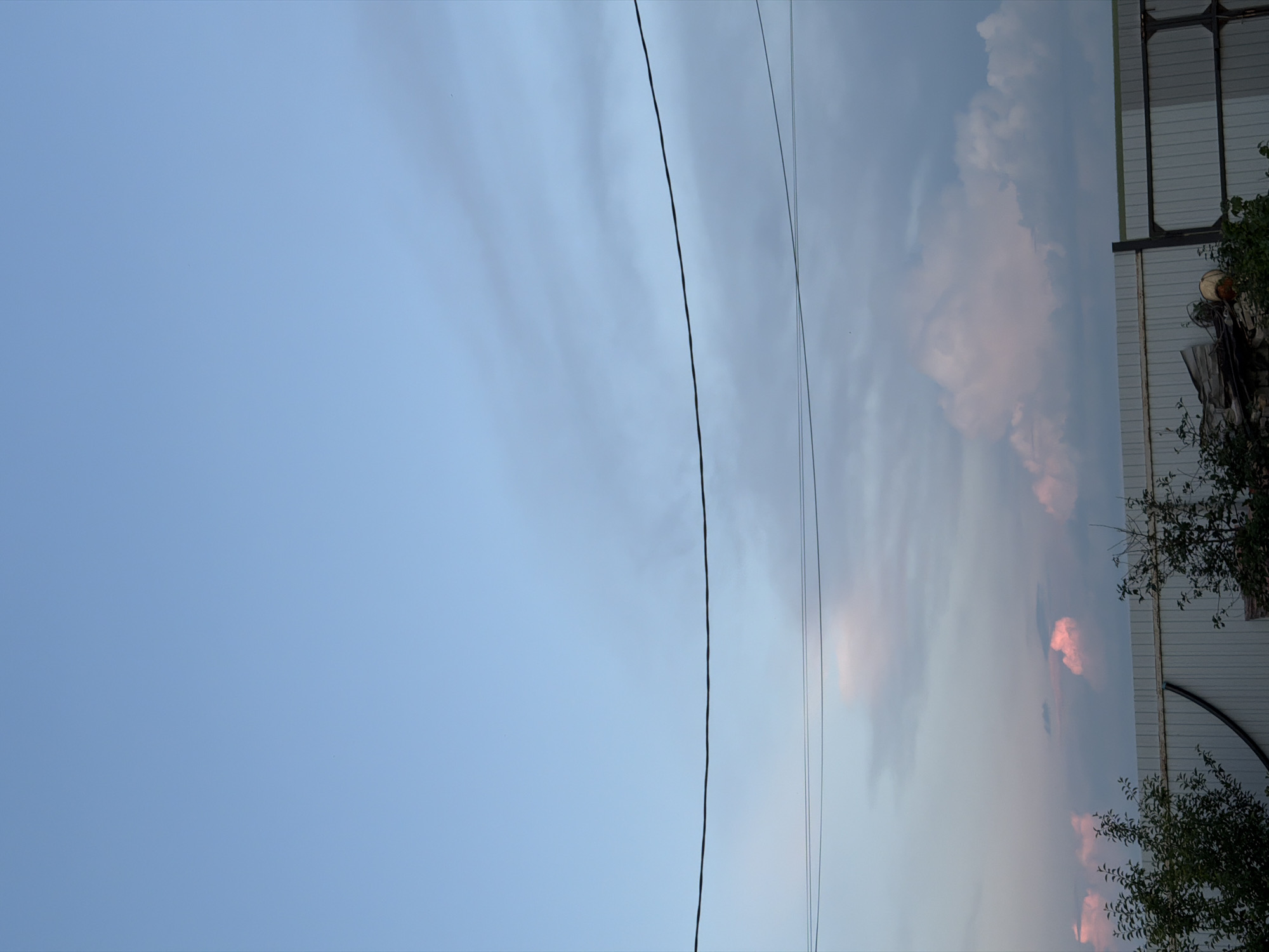 A low-angle, medium shot captures a sky with soft, pale blue hues gradually transitioning into gray clouds towards the horizon. Two distinct power lines stretch horizontally across the frame, creating a dividing line between the upper and lower portions of the image. Below the power lines, the sky is filled with scattered, fluffy clouds, some tinged with a soft pink or white. The bottom of the image features a white, corrugated metal fence running horizontally. On the left side of the fence, dark green foliage from trees and bushes partially obscures the fence. On the right side, the fence continues, with a dark metal gate or extension visible. There are no people or animals present in the scene. The overall environment appears to be an outdoor, possibly semi-rural or industrial, setting in Nizhniy Suyuk-Su, Russia, during twilight or early dawn, given the soft, diffused light and cloud coloration.