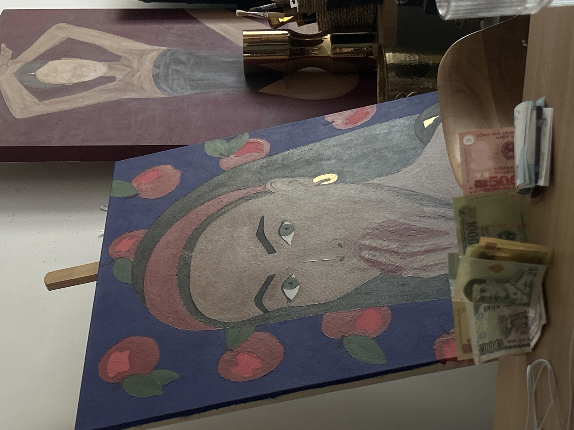The image presents two paintings, one depicting a figure in a reclining pose and the other featuring a portrait of a woman against a blue background with red floral motifs. A stack of Indonesian currency is visible to the right of the portrait painting. The scene appears to be set indoors, possibly in an art studio or gallery in North Kuta, Indonesia. The lighting in the image is diffused.