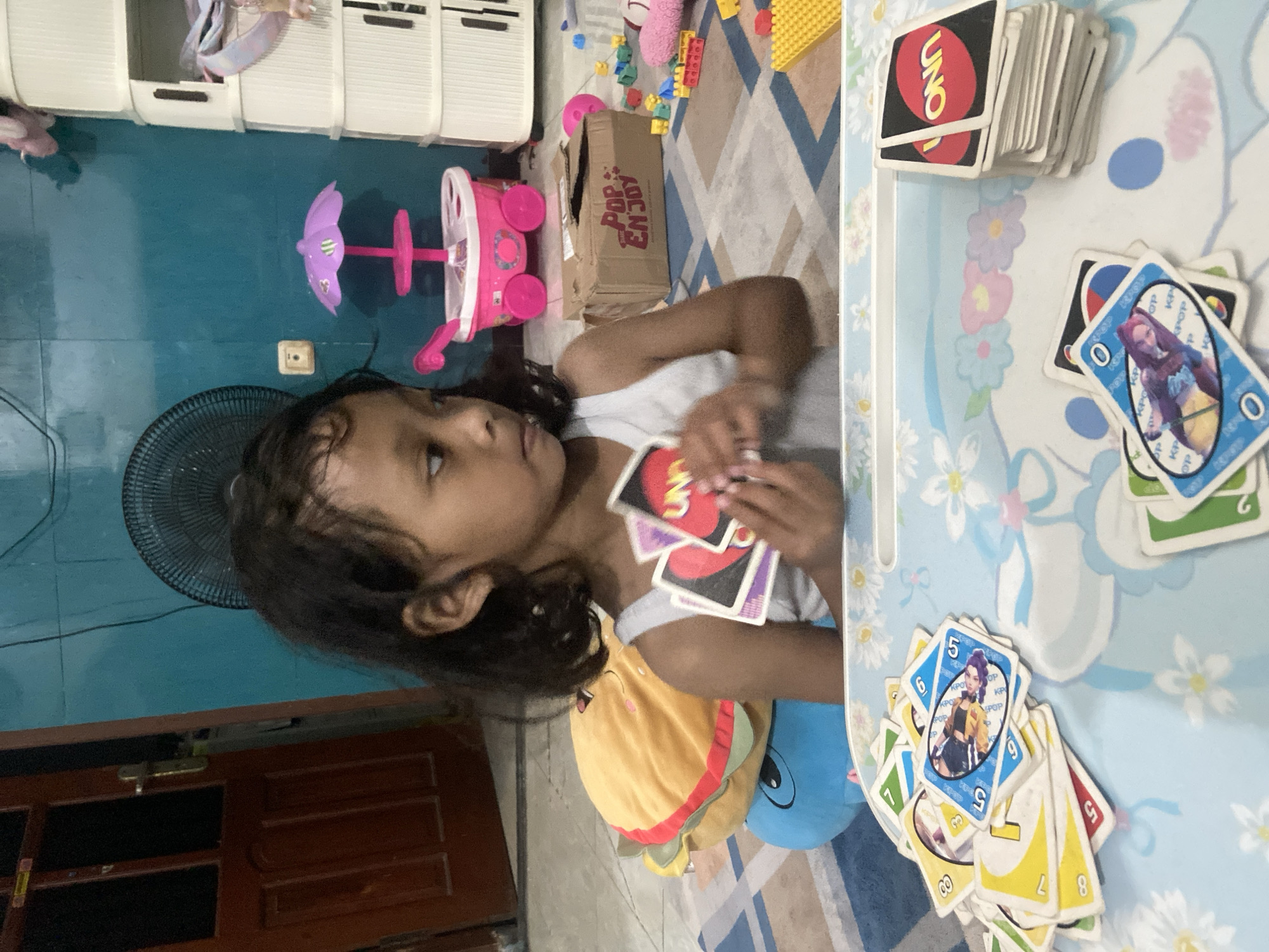 In Gelam, Indonesia, a young Indonesian girl with dark hair is holding a hand of UNO cards. She is wearing a white tank top and sitting at a light blue tray table with a floral pattern. A stack of UNO cards is on the right side of the tray, and several more UNO cards are scattered on the tray and in her hand. Behind the girl, a dark blue and yellow stuffed toy burger is visible. The room has a teal-colored wall and a fan in the background. To the right of the fan, a pink toy stroller with a purple umbrella is present. Further to the right, a stack of white drawers and a brown cardboard box with "POP ENJOY" printed on it are visible. The floor is covered with a blue and white patterned rug, with scattered colorful building blocks. The overall scene depicts a casual indoor setting where the girl is engaged in a card game.