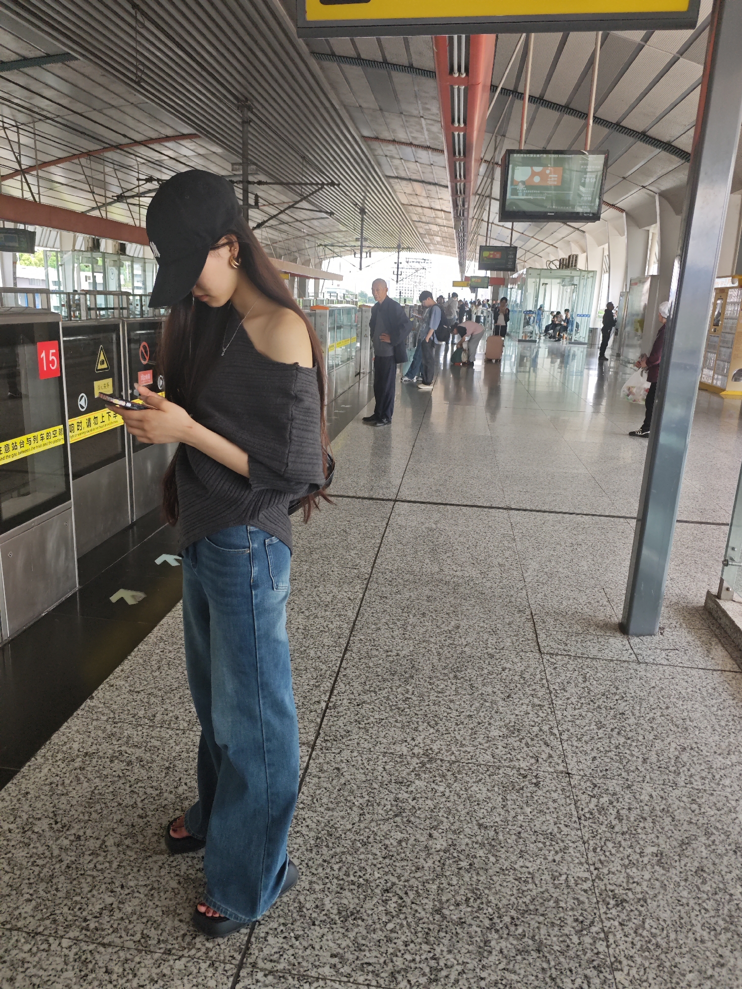 The image shows a young woman standing at a train station platform, likely waiting for a train. She is looking at her phone and wearing a black baseball cap, a gray off-the-shoulder sweater, loose blue jeans, and black platform sandals.
The platform is clean and well-lit with a speckled gray floor. Behind her is a platform door with a sign with a warning in Chinese, and yellow safety tape. There are other people in the background, some standing and waiting, others walking. Above, there are metal beams and a television screen.
Based on the location information, this image was likely taken in the Chongqing High-tech Zone, China.