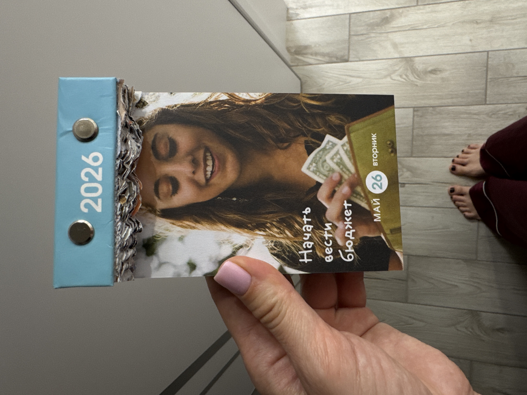 A calendar held by a hand, with a woman on the cover holding cash, displays the date "May 26" and the year "2026." The calendar's blue binding is visible, along with metal rings. In the background, a glimpse of a person's feet with painted toenails is seen on a light-colored, wood-patterned floor. Part of a gray wall or cabinet is also visible on the left. There is no indication that the picture was taken in Sochi, Russia.