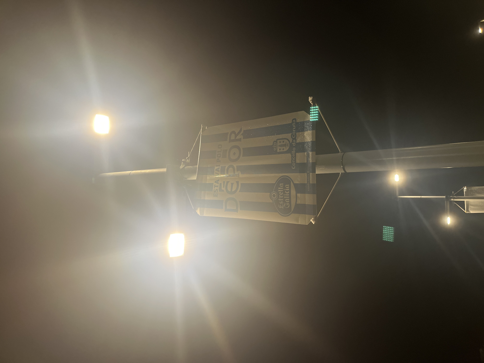 A vertical view depicts a lamppost at night in A Coruña, Spain. The lamppost is topped with a sign featuring blue and white stripes and the word "DEPOR," partially obscured by glare. The sign also displays the "Estrella Galicia" logo and mentions "Concello da Coruña." The surrounding environment is dark, with illumination primarily from the lamppost's light and stadium lights.