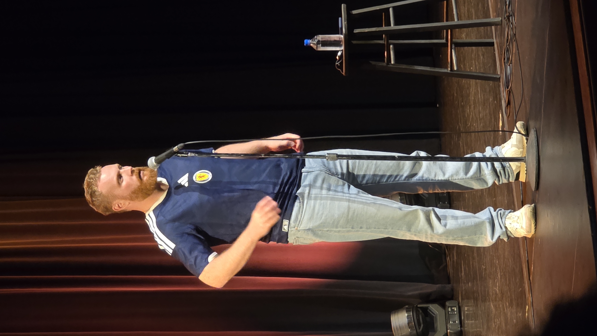 A man with red hair and a beard is standing on a stage in front of a microphone. He is wearing a dark blue soccer jersey with white stripes on the sleeves and a crest on the chest, light blue jeans, and light-colored sneakers. His left arm is bent at the elbow and his hand is near his hip, while his right arm is extended forward. Behind him, red curtains are visible on the left side of the stage, and a black backdrop is on the right. To the right of the man, a dark stool with a water bottle on it is positioned on the stage. Stage lights are visible on the floor to the left of the man. The man appears to be speaking or performing. The location is the Comedy Underground in Seattle, Washington, United States of America.