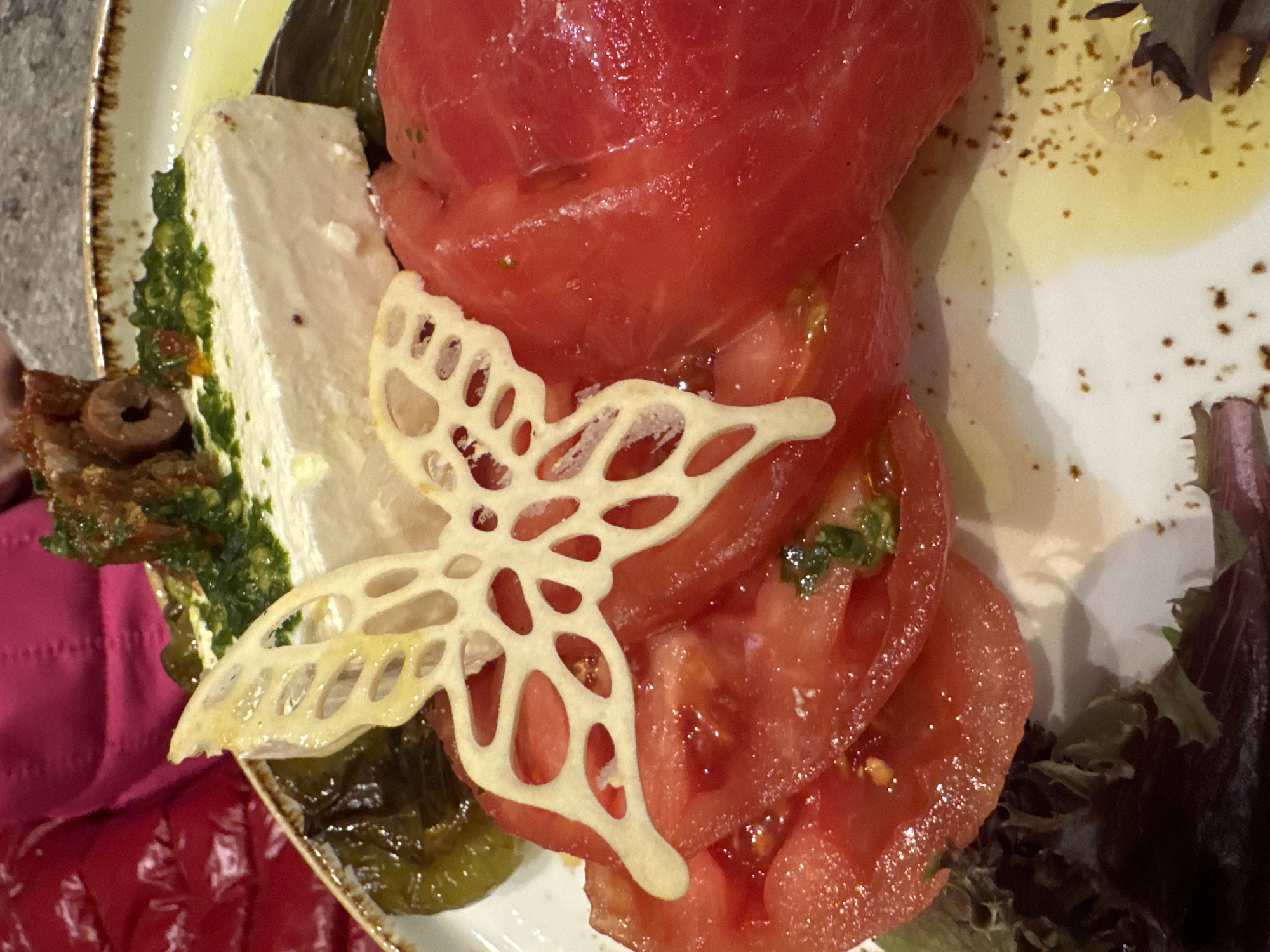 The image shows a plate of food, possibly in a restaurant. The plate contains sliced tomatoes stacked on top of each other. There is a white, rectangular block next to the tomatoes, possibly cheese with a green pesto-like sauce. A decorative butterfly-shaped garnish is placed on the tomatoes. There are also olives and mixed greens on the plate. Some oily dressing or sauce is visible on the plate. The location cannot be determined from the image.