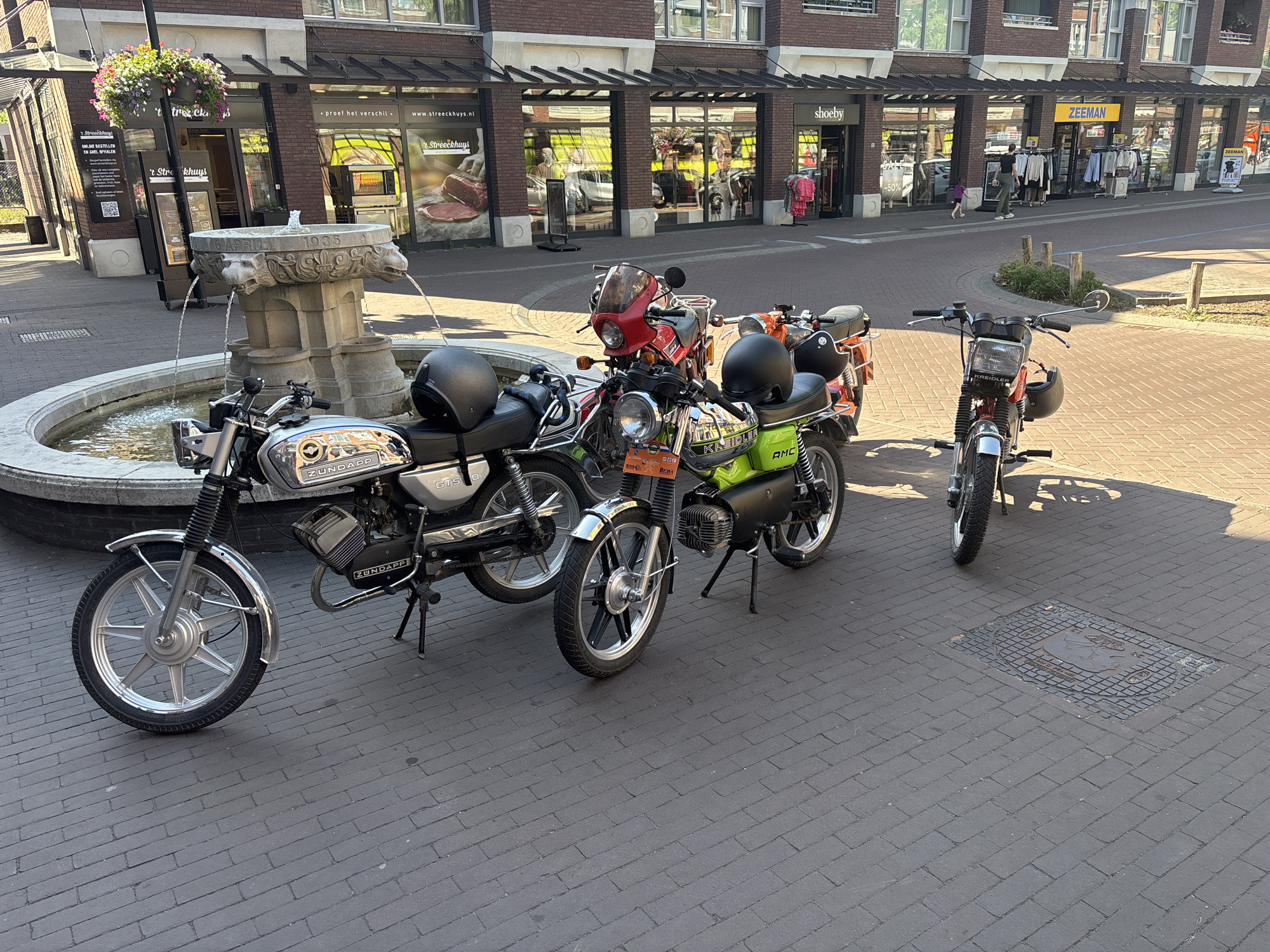 The image depicts a street scene in the city of Asten, Netherlands. In the foreground, three vintage motorcycles are parked on a brick-paved sidewalk. The motorcycle on the left is silver and black with "ZUNDAPP" branding. The middle motorcycle is bright green with black accents and a "KREIDLER" logo. The motorcycle on the right is red with a black seat and a small windshield. Two helmets are visible, one on the silver motorcycle and another on the green motorcycle. A stone fountain with water flowing from it is situated behind the motorcycles. In the background, a brick building with several storefronts is visible. Storefronts include "shoeby" and "ZEEMAN". People are seen walking in front of the stores. The overall environment suggests a sunny day in a commercial area.