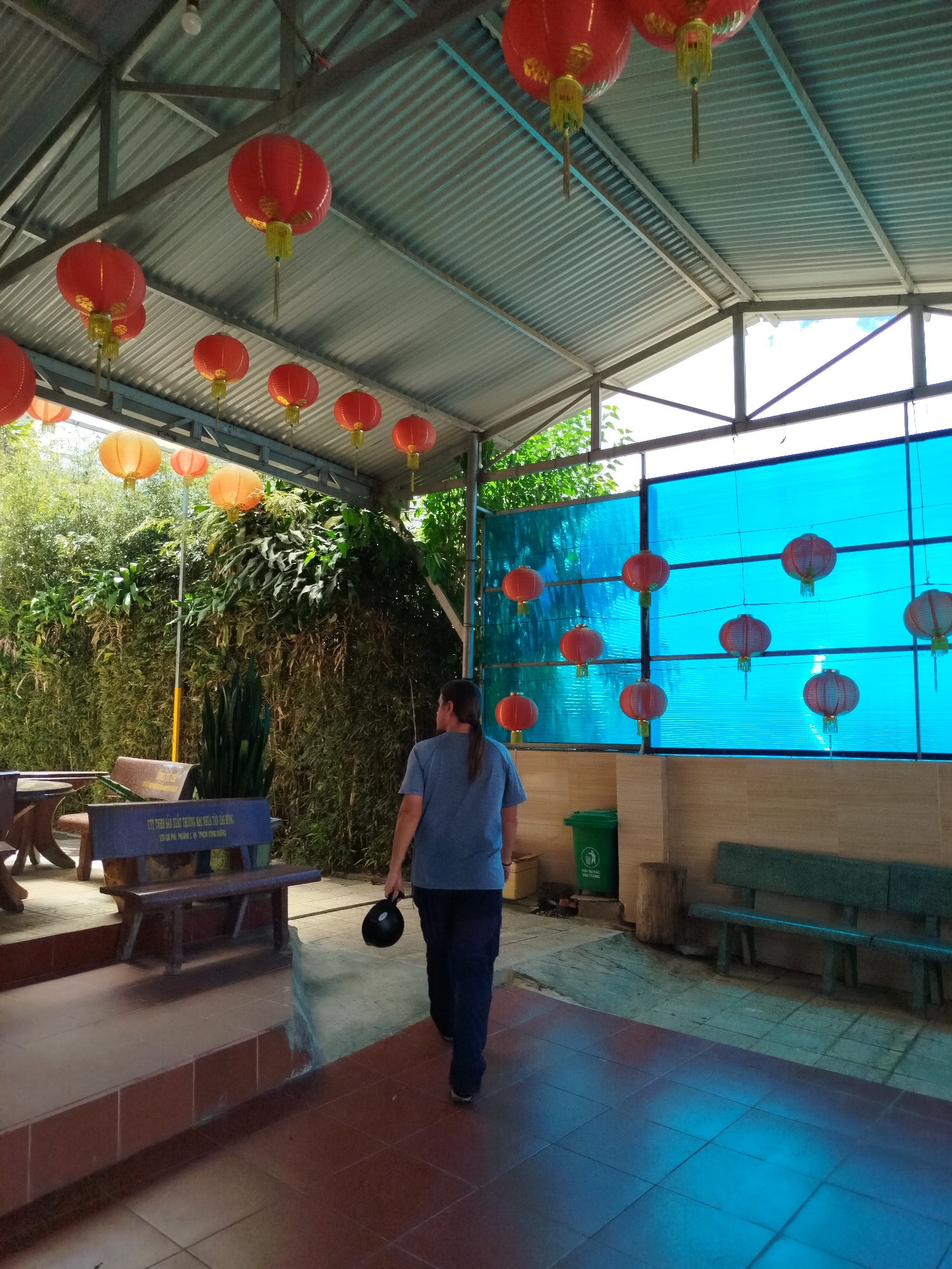 A person walks through a covered area decorated with red and orange lanterns in what appears to be Da Lat, Vietnam. The person, seen from the back, is wearing a gray shirt and dark pants, and carrying a black object in their left hand. The covered area has a corrugated metal roof supported by a metal frame. Red lanterns hang from the roof, and in the background, a blue translucent panel is adorned with more red lanterns. To the left, a bench with text in Vietnamese sits on a raised platform next to tables and dense green vegetation. To the right, a bench sits against a tiled wall, with a green trash bin nearby. The floor is tiled.