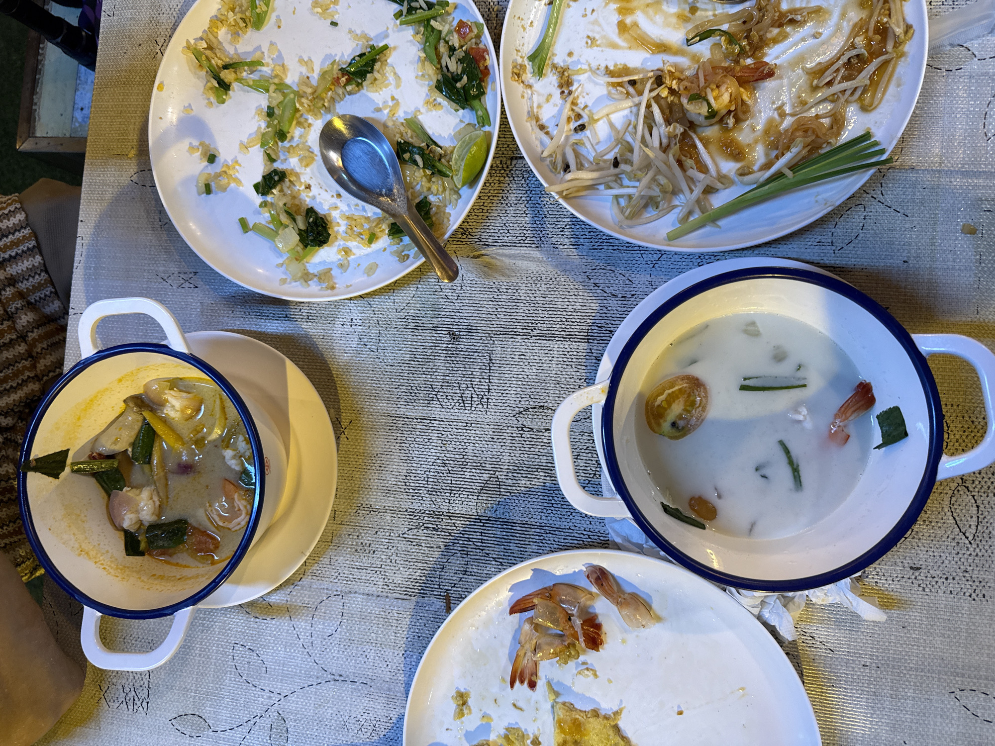 The image shows a table with several plates of partially eaten food. In the center of the frame, a large white bowl with a blue rim contains a creamy soup with chunks of meat, vegetables, and what appear to be pieces of shrimp. To the right of this bowl is a white plate with remnants of fried rice and some greens, with a silver spoon resting on it. Below this plate is another white plate with a portion of Pad Thai, including bean sprouts, noodles, and pieces of shrimp. To the left of the central soup bowl is a white plate with a few pieces of shrimp and some yellow material, possibly egg. A third white bowl with a blue rim is visible at the bottom center, containing a lighter colored soup with pieces of tomato and shrimp. The table is covered with a textured cloth. The lighting suggests an indoor or outdoor evening setting. No people are clearly visible in the image.