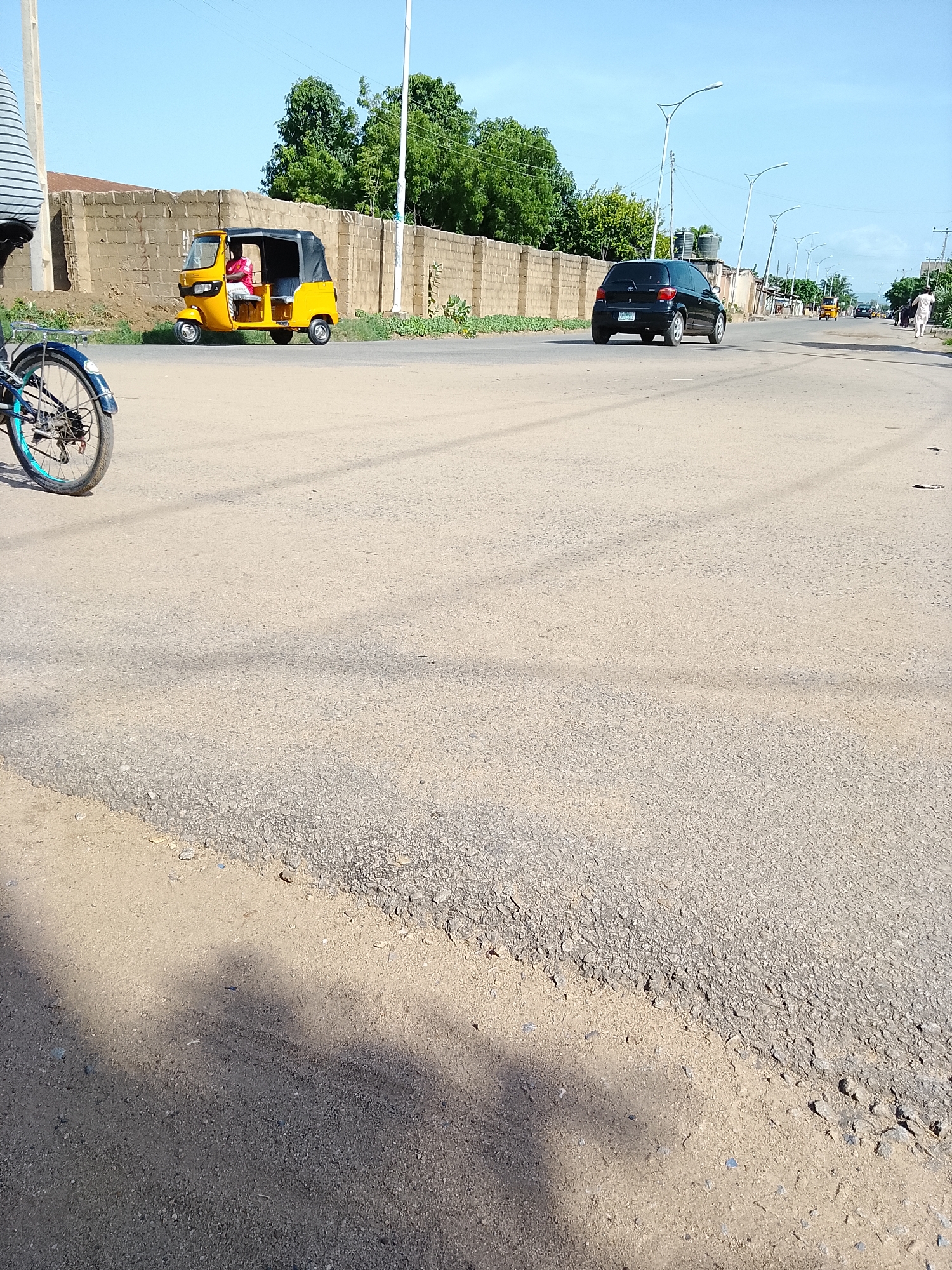 In Wauro Leggal, Nigeria, a yellow three-wheeled auto-rickshaw, driven by an individual wearing a pink shirt, is traveling on a paved road. The auto-rickshaw has a black fabric roof. Adjacent to the road is a low, brick wall. In the background, a black sedan is visible, and beyond that, streetlights line the road. Trees are present behind the wall and in the distance. The sky is clear and blue, indicating a sunny day. The road surface is composed of asphalt. A bicycle with a blue rim is partially visible on the left side of the image, its wheel closest to the viewer. The scene appears to be an ordinary street in a town.