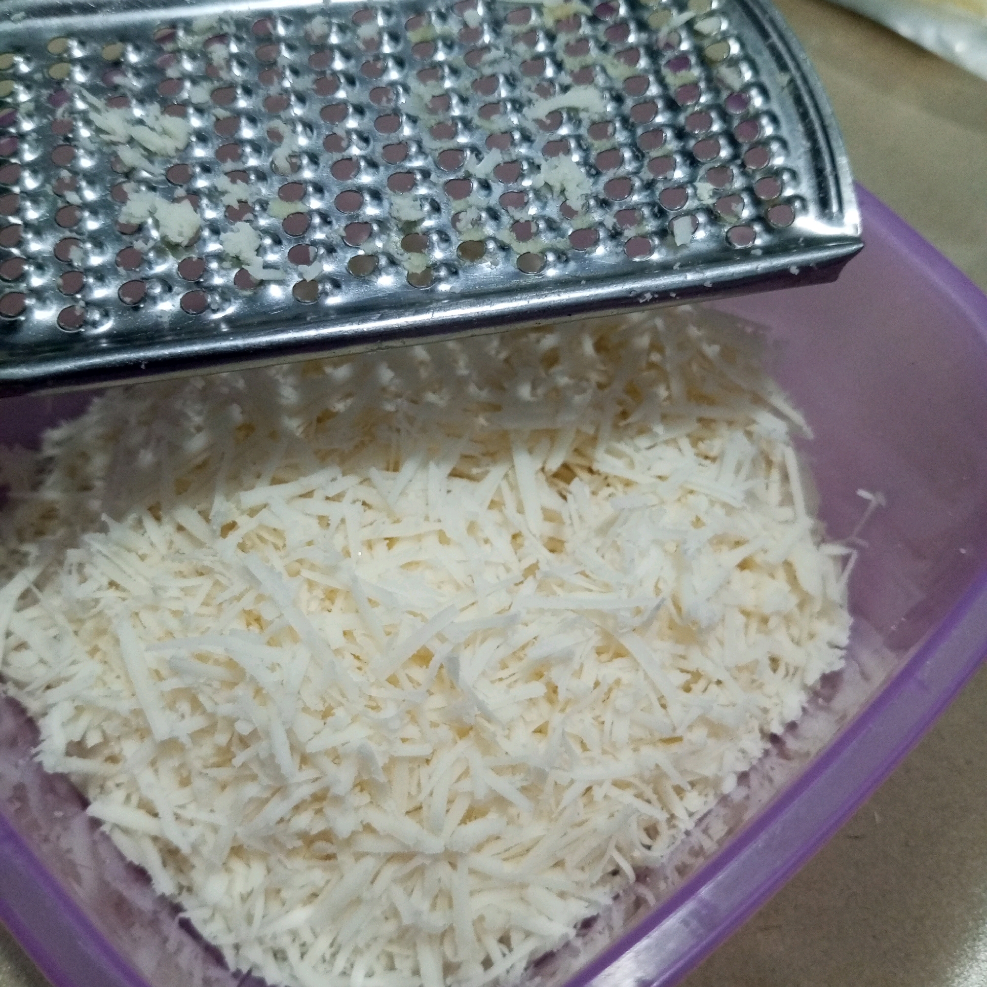 A close-up, top-down shot shows grated cheese being prepared. A metal grater with many small, circular holes rests on the edge of a translucent purple plastic container. Some cheese shreds are visible on the grater, while a large pile of finely grated white cheese fills the container. The background is a neutral surface, possibly a countertop. The lighting is even, highlighting the texture of the cheese and the metallic sheen of the grater. There are no people or indications of the specific location in North Jakarta, Indonesia, visible in this image.