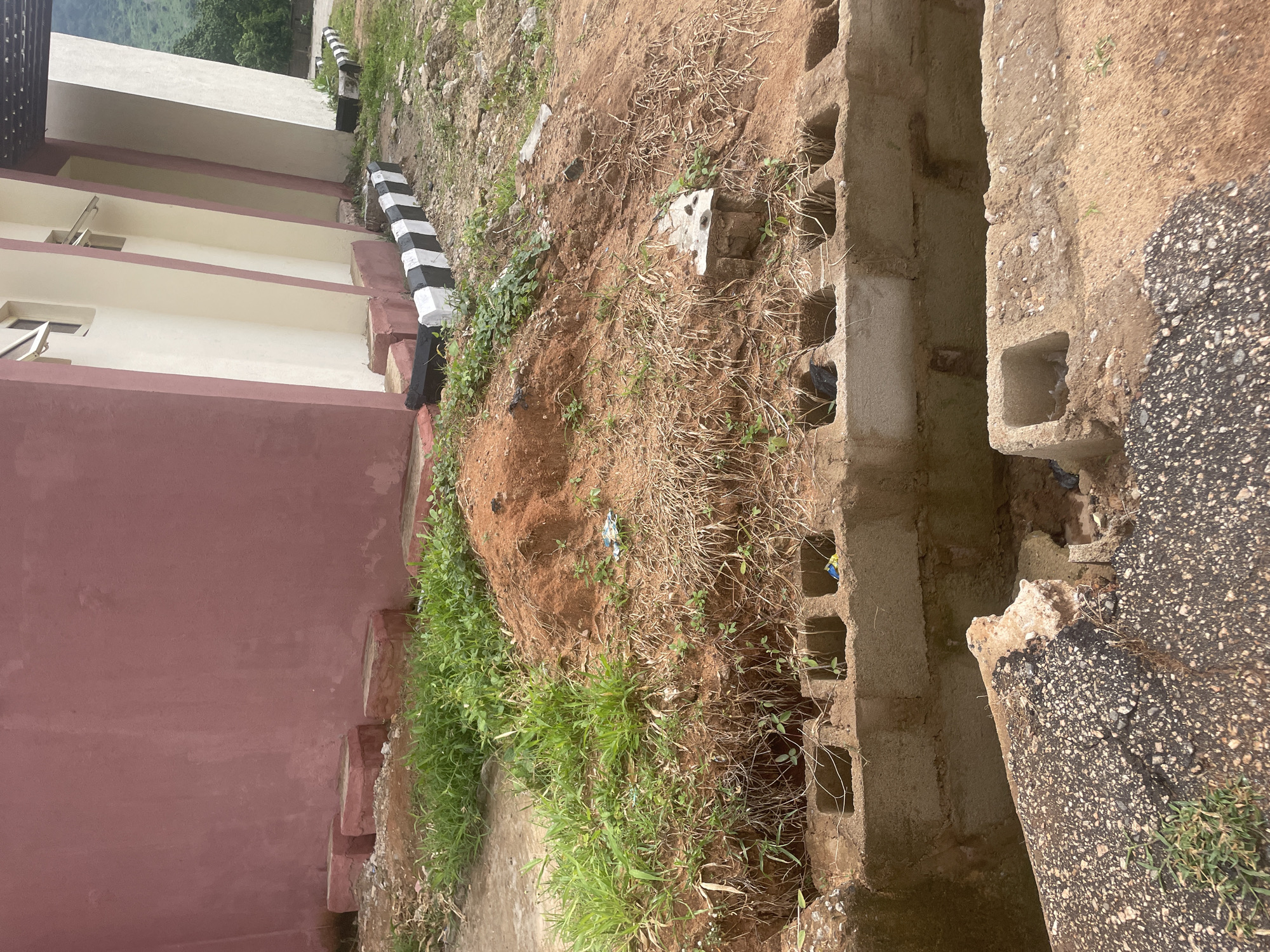In Lokoja, Nigeria, this image captures a section of an unfinished or damaged construction site. A muted pink building with cream-colored columns and black and white striped barriers is visible in the background. In the foreground, a mound of reddish-brown soil with sparse green and dry vegetation sits atop a foundation constructed from hollow concrete blocks. A broken asphalt surface is also present in the lower portion of the frame, partially obscuring more concrete blocks. The scene appears to be outdoors in an area that is either under construction or has fallen into disrepair.
