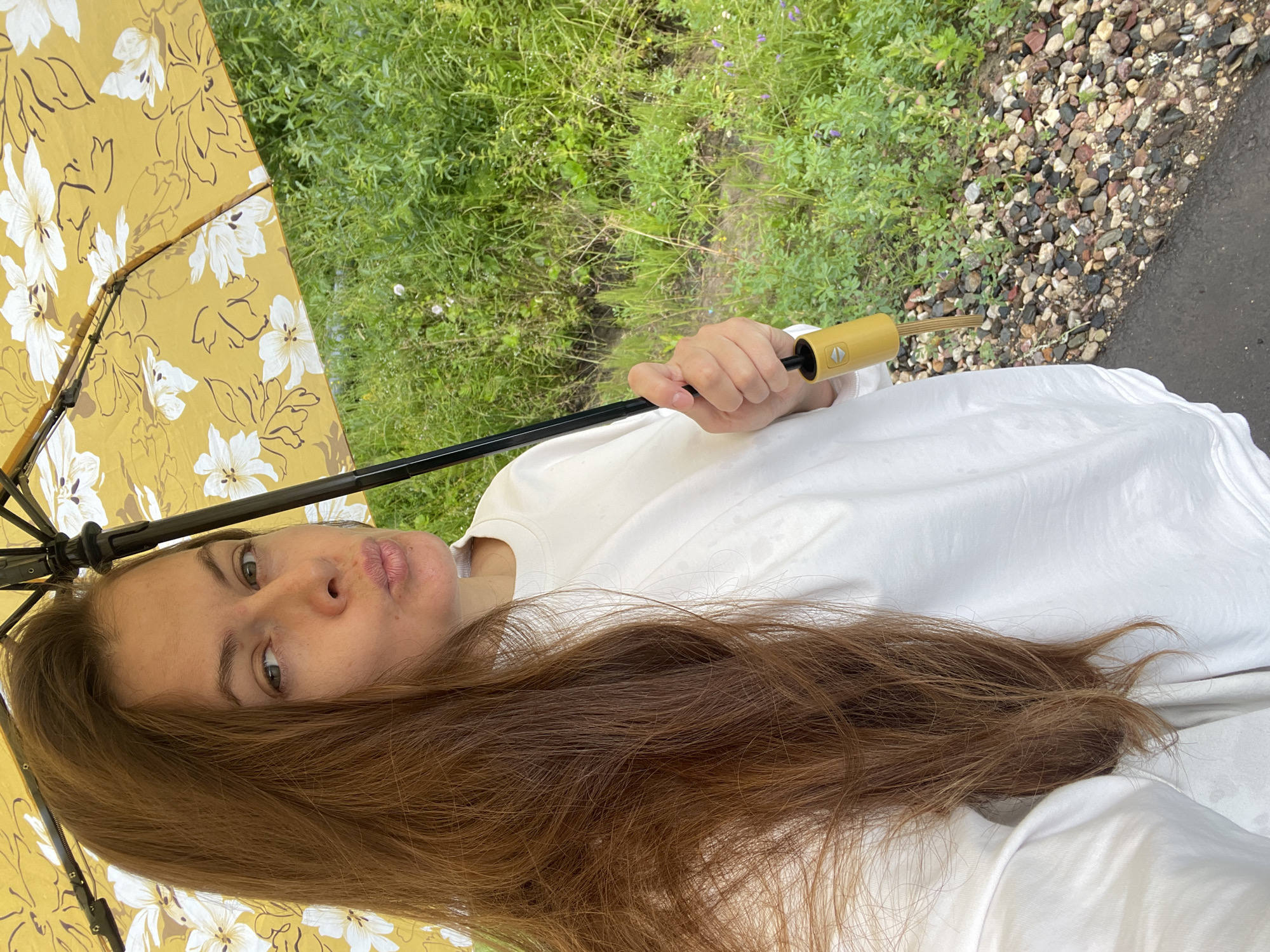 In this close-up, vertically oriented selfie, a person is holding an open yellow umbrella with a white floral pattern. The umbrella is positioned above and to the left of the subject, partially obscuring their face and hair. The person, seen from the chest up, has long, auburn hair that falls over their shoulder and onto a white t-shirt. Their face is turned towards the camera, with eyes looking slightly to the side and lips pursed.
The background reveals a natural setting. To the left and behind the umbrella, lush green grass and vegetation are visible, dotted with small purple wildflowers. To the right, a path or area covered in various-sized, multi-colored stones leads away from the viewer, bordered by what appears to be a wet, dark surface, possibly asphalt. The lighting suggests it is daytime, and the presence of the umbrella hints at overcast or rainy weather. The overall mood is casual and perhaps a bit whimsical due to the floral umbrella. No visible text is present in the image. The location context is Заречный, Russia.
