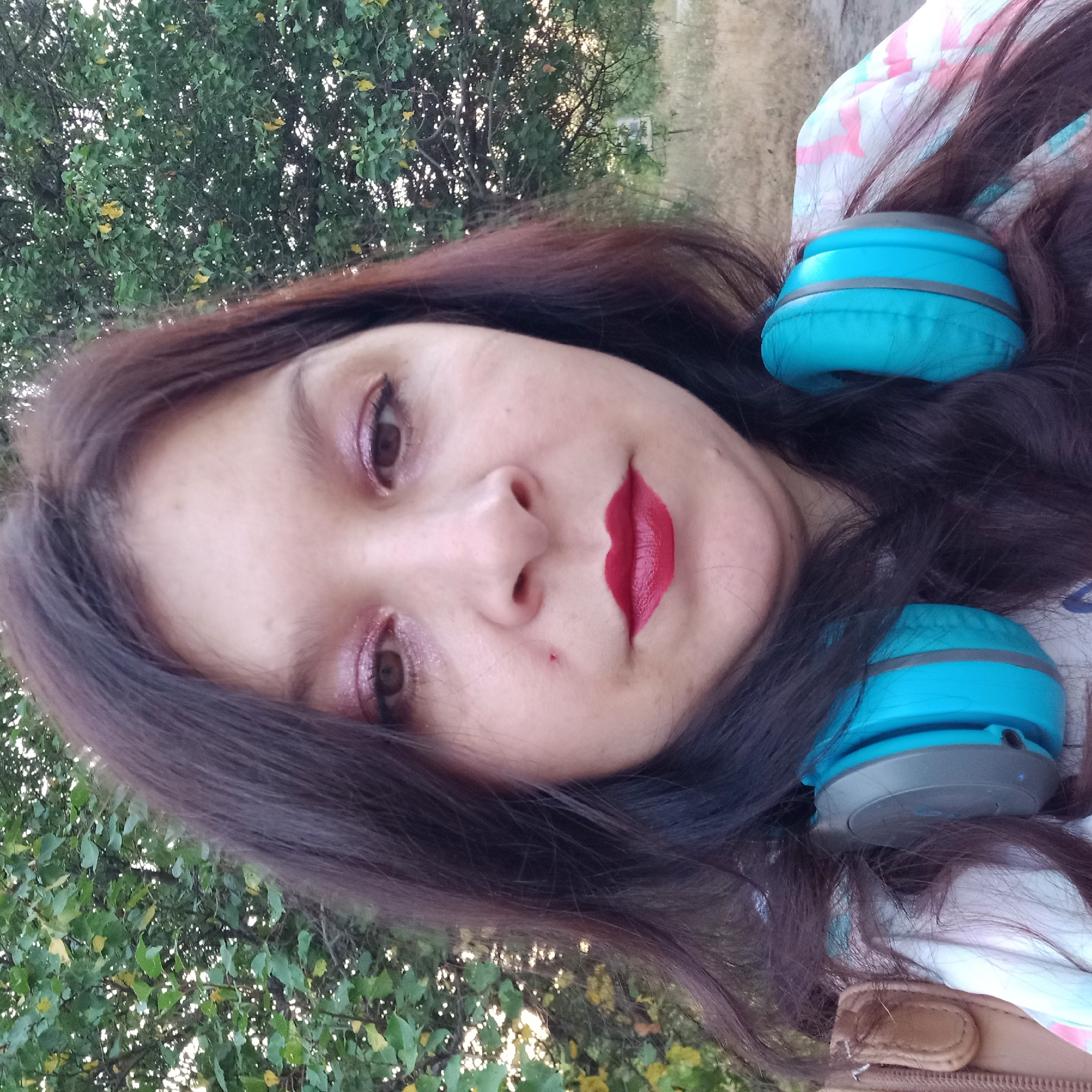 A woman with long, dark hair and prominent makeup, including shimmery pink eyeshadow and red lipstick, is pictured from the chest up in an outdoor setting. She is wearing bright turquoise headphones around her neck. Behind her, a dense green tree with some yellowing leaves is visible, suggesting an outdoor environment possibly in Neopalymivka, Ukraine during the autumn season. The lighting indicates it is daytime, possibly late afternoon given the soft light filtering through the leaves. She is looking directly at the camera.
