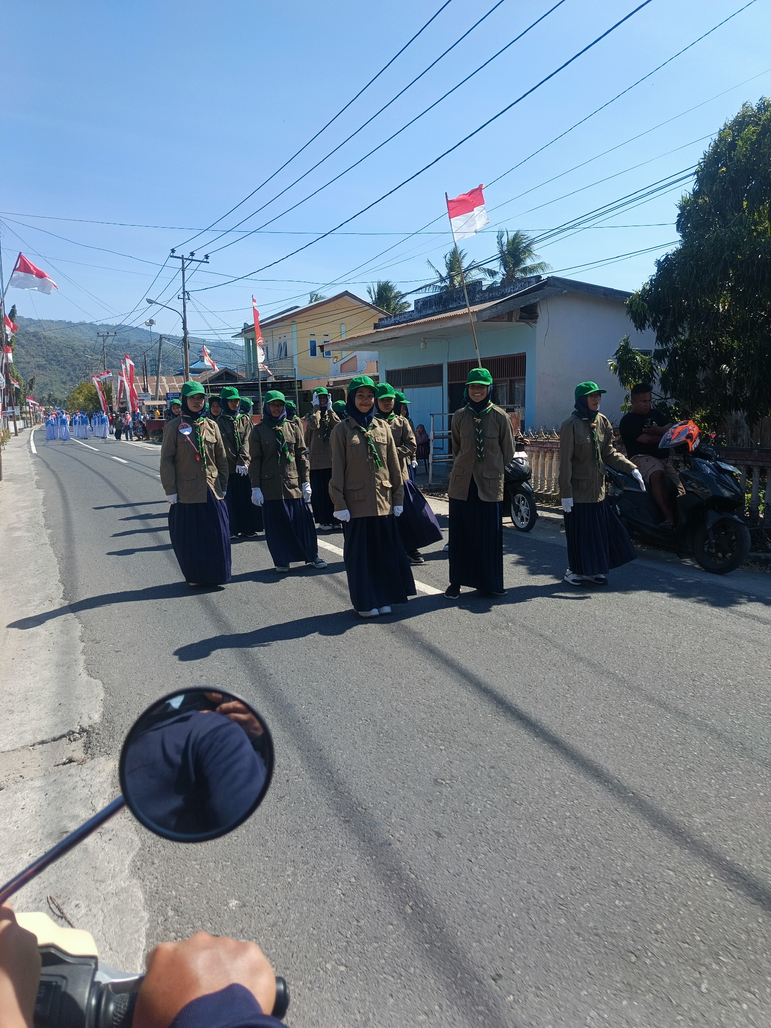 In Tenggela, Indonesia, a group of young people in scout uniforms, wearing green caps, brown jackets, and dark blue skirts, participate in a parade on a paved road under a clear blue sky. Indonesian flags are visible on poles lining the street, and several buildings are present in the background, along with trees and utility lines. The perspective is from someone riding a motorcycle, with a mirror reflecting the rider and the handlebars. Other people on motorcycles and a distant group in white uniforms are also part of the scene. The overall atmosphere suggests a celebratory or ceremonial event.
