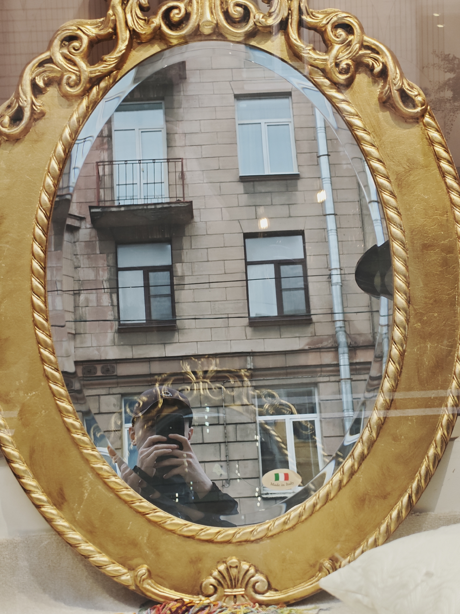 A mirror with an ornate gold frame reflects a building with multiple windows and a small balcony. In the reflection, a person is taking a picture of themselves with a smartphone. The person is wearing a dark cap and jacket. A small oval sticker with the Italian flag and the words "Made in Italy" is visible on the glass. The background suggests an urban environment with a brick building and visible pipes.
