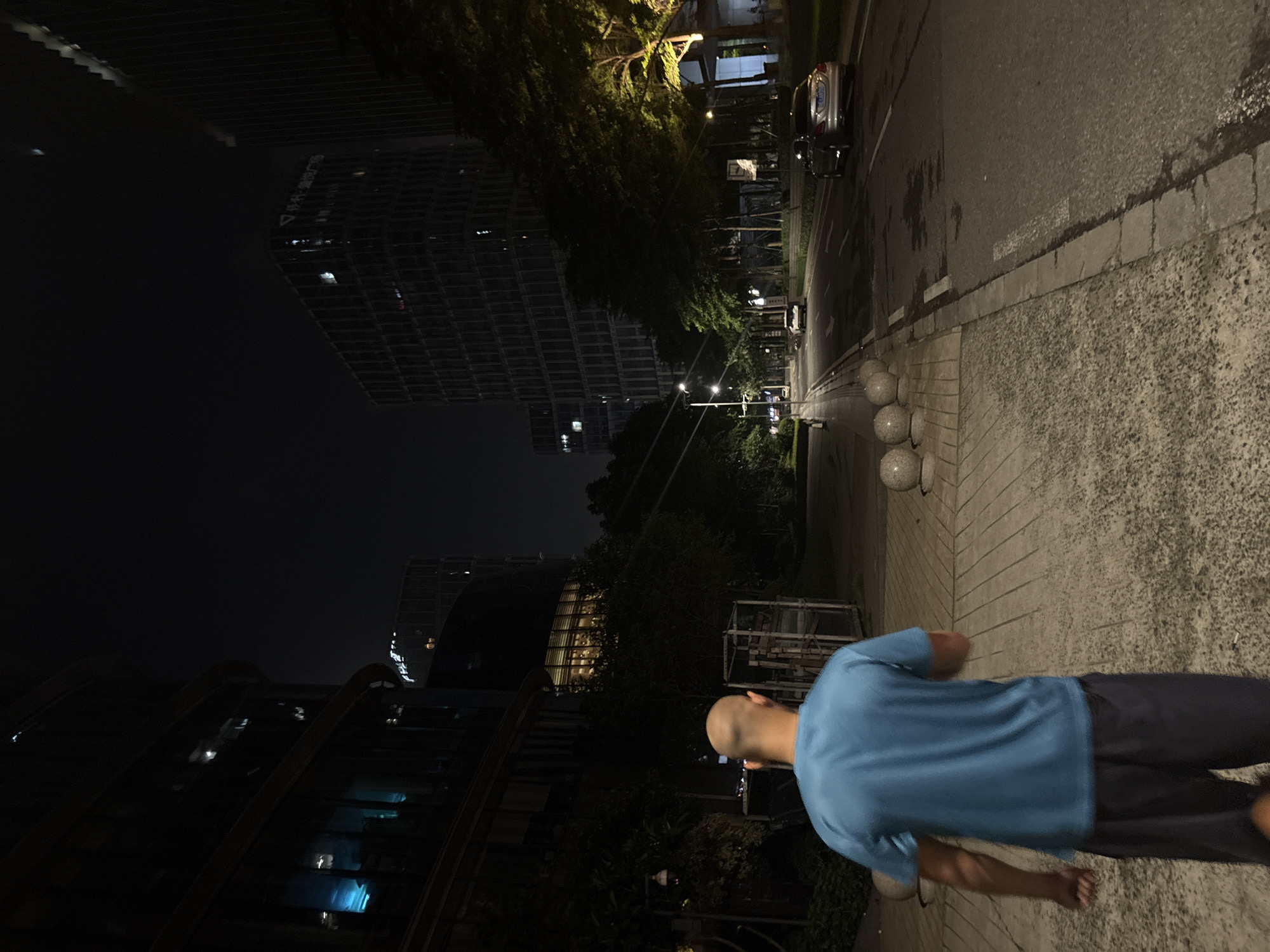 The image depicts a nighttime scene in Haizhu District, China. The primary subject is a bald man from behind, wearing a blue t-shirt and dark shorts, standing on a paved walkway. The man is facing away from the camera, towards a street with a car and streetlights. In the background, large, modern buildings with illuminated windows are visible, suggesting an urban environment. The lighting indicates it is nighttime. The overall mood appears calm and perhaps contemplative, with the man standing alone amidst the city. The paving of the walkway is made of rectangular blocks, and the street is asphalt with white markings. There are also decorative spherical bollards along the edge of the walkway.