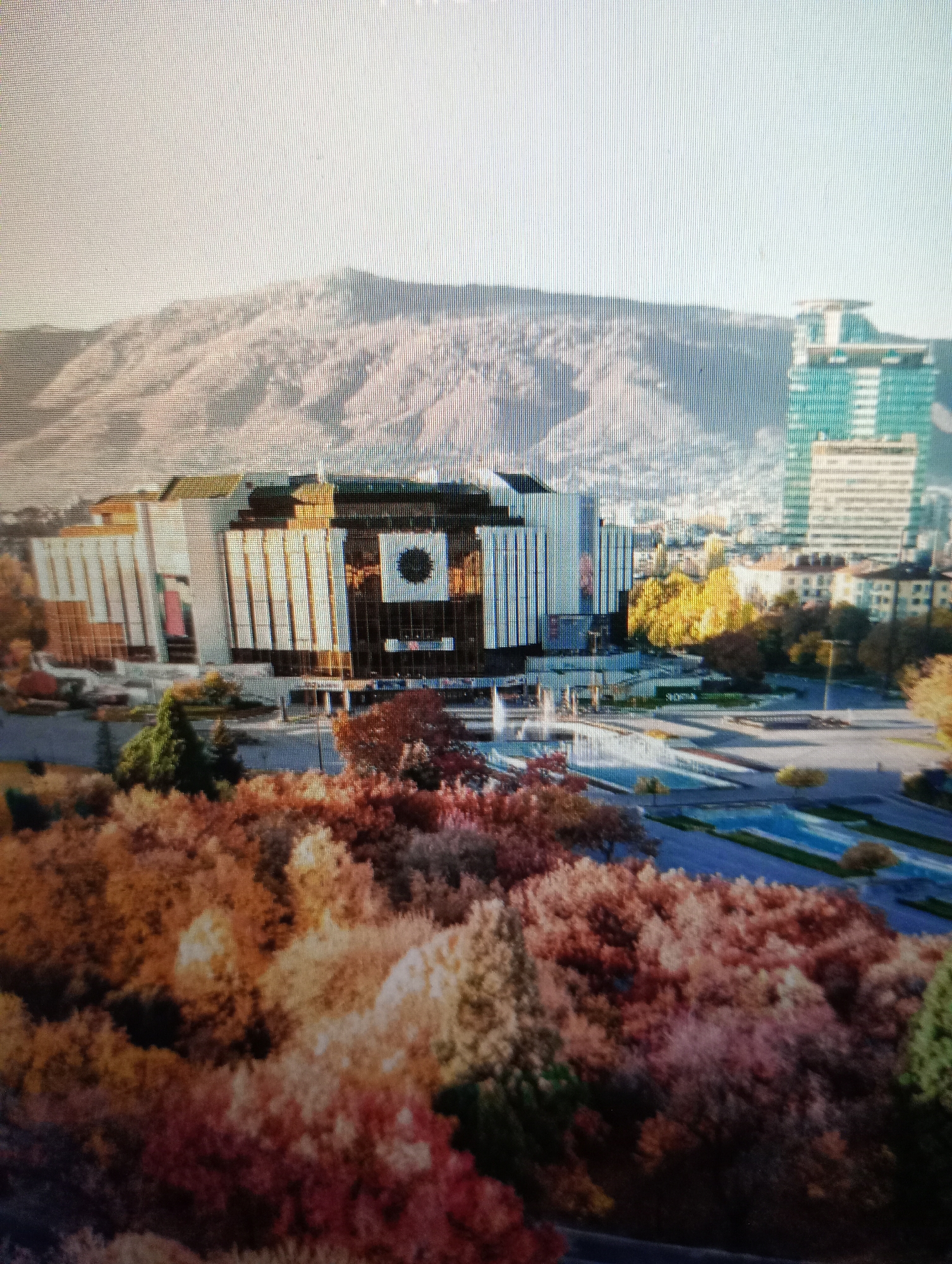 The image shows an aerial view of the National Palace of Culture in Sofia, Bulgaria, with a focus on its facade and the surrounding parkland. The National Palace of Culture is a large, multi-purpose complex with a distinct architectural style, characterized by its imposing structure and large glass panels. In the foreground, a dense canopy of trees exhibits the vibrant colors of autumn, with hues of red, orange, and yellow dominating the scene. Behind the palace, a modern skyscraper, possibly the Interpred World Trade Centre, stands tall against the backdrop of a mountain range. The sky is clear and bright, suggesting a sunny day. The image is a static depiction of an urban landscape in Sofia, capturing the interplay between natural elements and architectural structures.