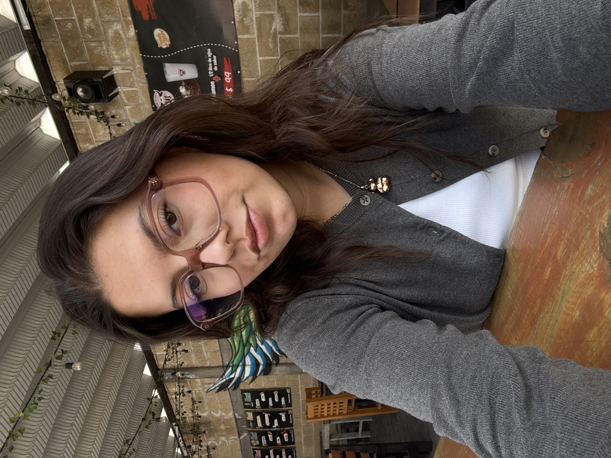 The image is a vertical selfie of a young woman with long, dark brown hair and light pink-framed glasses, looking directly at the camera with a neutral expression and slightly parted lips. She is wearing a charcoal grey button-up cardigan over a white top, with a delicate gold-toned necklace featuring a small, ornate golden charm around her neck.
She is seated at a rustic wooden table with visible wear and tear. The setting appears to be an outdoor or semi-outdoor cafe or restaurant, suggested by the exposed brick wall in the background and the natural lighting. On the brick wall, a black speaker is mounted, and string lights with Edison-style bulbs are strung along with some green foliage.
A black poster or menu board is visible on the brick wall behind her, displaying white text. Partially legible text includes "1/2 litro de agua de sabor" (1/2 liter of flavored water) and "+ $99". The word "ramen" is also partly visible. Below this, there are smaller posters with unreadable text and images.
In the upper background, a light-colored, possibly corrugated or stucco ceiling is visible, adorned with more string lights. To her left, a painted mural of a wing with blue, green, and white feathers can be seen. Further back, wooden tables and chairs indicate a dining area. The lighting suggests it is daytime. The location context is San Andrés Cholula, Mexico.