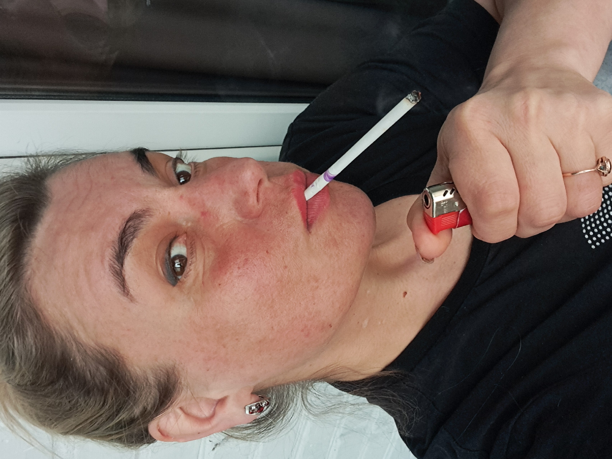 A woman is shown from the chest up, holding a lit cigarette in her mouth and a red lighter in her right hand. She is wearing a black t-shirt, and a ring on the ring finger of her right hand. She has earrings and light gray hair. The location is unknown and no element indicates that it is in the city of Elektrostal, Russia.