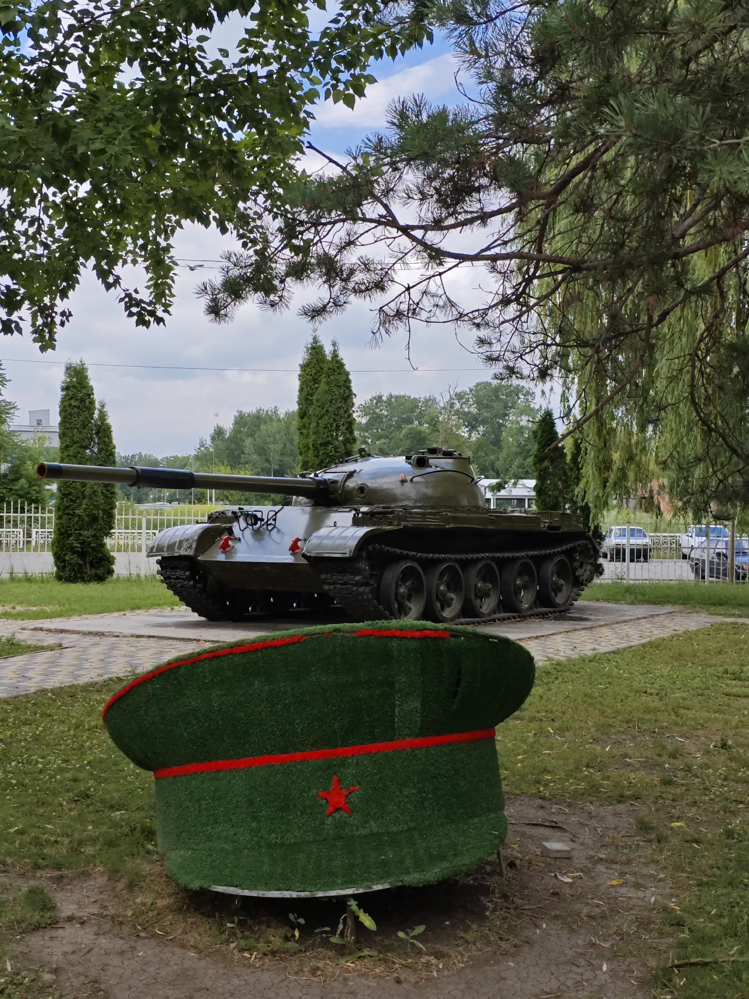 The image depicts an outdoor scene in Cherkessk, Russia, featuring a decommissioned military tank as a monument. In the foreground, a large, artificial green cap with a red band and a red star is prominently displayed. Behind the cap, the tank is positioned on a paved area surrounded by grass and trees. The tank is dark green, with visible tracks and a long barrel pointing to the left. Several tall, conical evergreen trees are planted behind the tank, and a white fence runs along the background. Beyond the fence, cars are visible parked on a street, and more trees form a dense backdrop. The sky is partly cloudy. The scene appears to be a public space, possibly a park or memorial site.