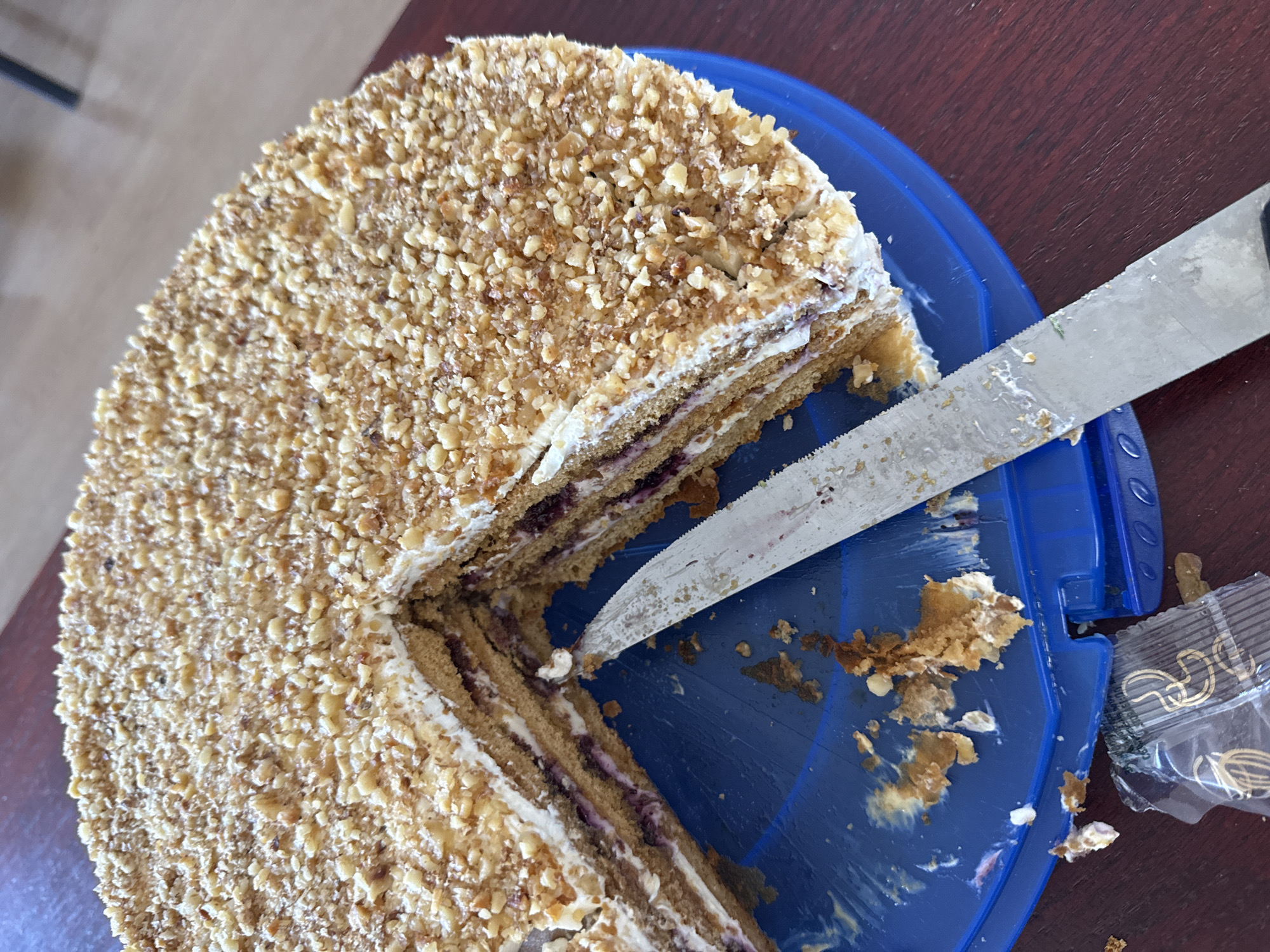 A close-up shot of a partially cut cake on a blue plastic plate. The cake appears to be multi-layered with a cream filling and a topping of finely chopped nuts. A serrated knife is partially inserted into the cake, indicating a slice has been removed. Crumbs and remnants of the cake are scattered around the cut portion on the plate. A sealed package of what appears to be paper clips or similar small metallic items is also visible on the plate's edge. The background surface is a dark wood grain. Location cannot be determined based on the image alone.