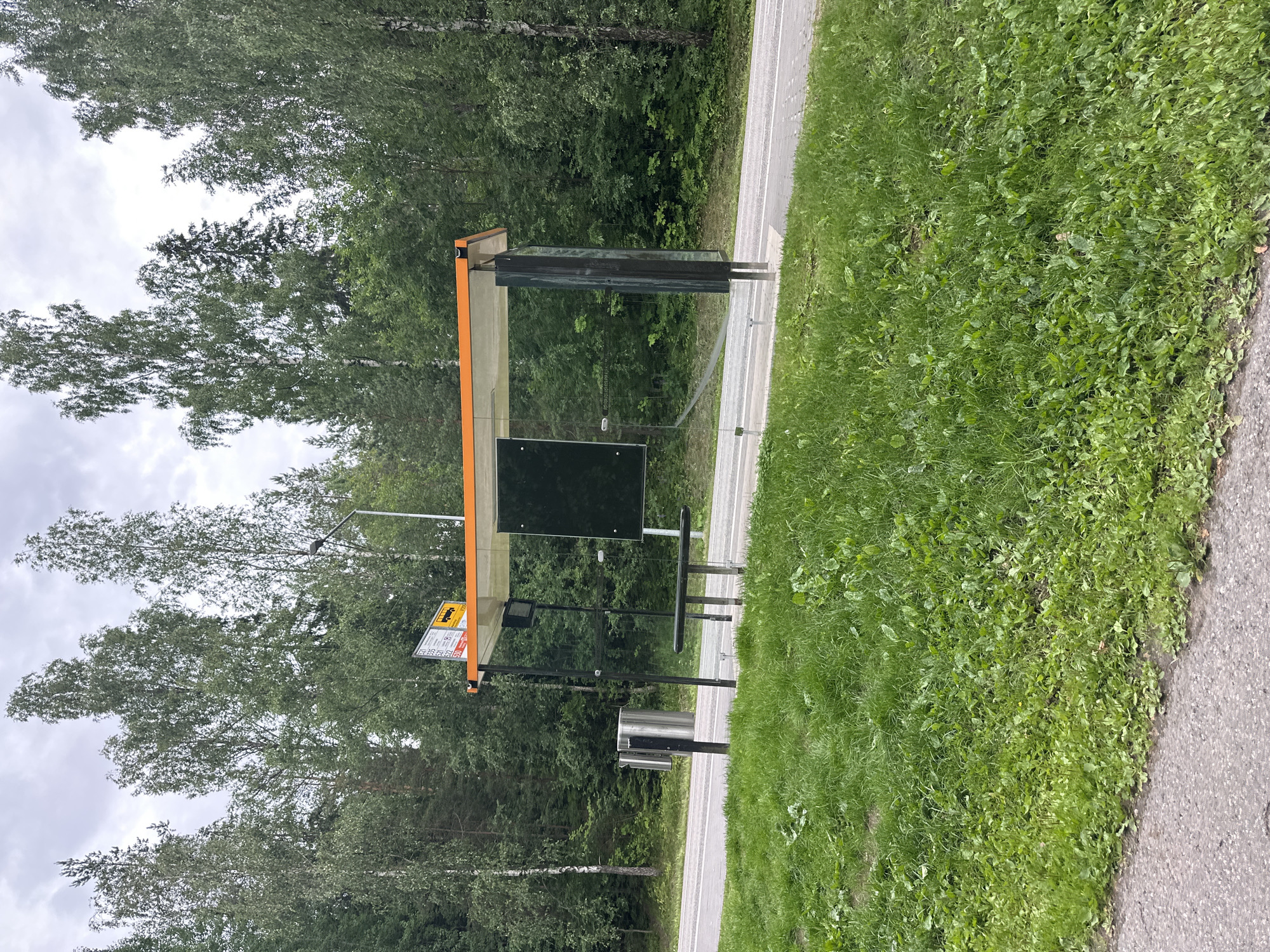 A bus stop shelter with an orange roof and clear glass panels is situated on the side of a paved road, surrounded by lush green grass and dense trees. The shelter has a black bench inside and a trash can made of metal next to it. A sign with bus route numbers and a yellow bus icon is attached to the top of the shelter. The sky above is overcast with clouds.