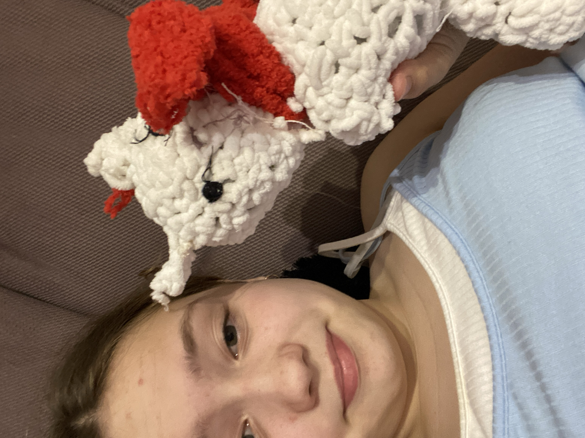 A young person is lying down indoors on a gray surface, possibly a couch or bed. The young person is holding a white, crocheted bear-like toy with red ears and black eyes above their head. The young person is wearing a light blue tank top with white trim. The specific location is unknown.