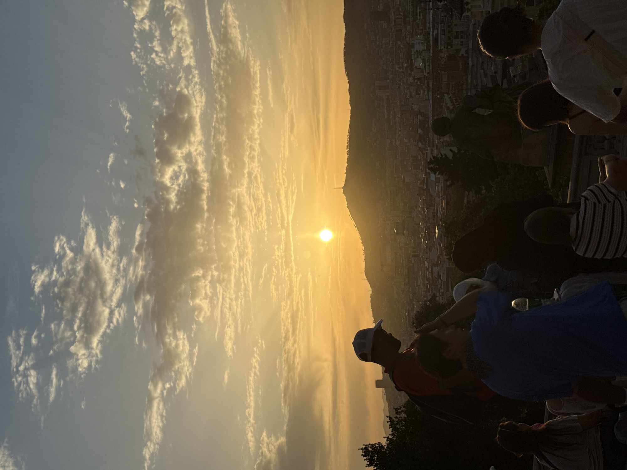 A group of people are gathered on a vantage point overlooking Barcelona, Spain at sunset. The sun is setting behind a distant hill, casting a warm glow over the city. The sky is filled with scattered clouds, illuminated by the setting sun. In the foreground, silhouetted figures of people are visible, some looking out at the view. The city of Barcelona stretches out below, with buildings and lights visible in the twilight. The overall scene captures a moment of observation and appreciation of the cityscape at dusk.